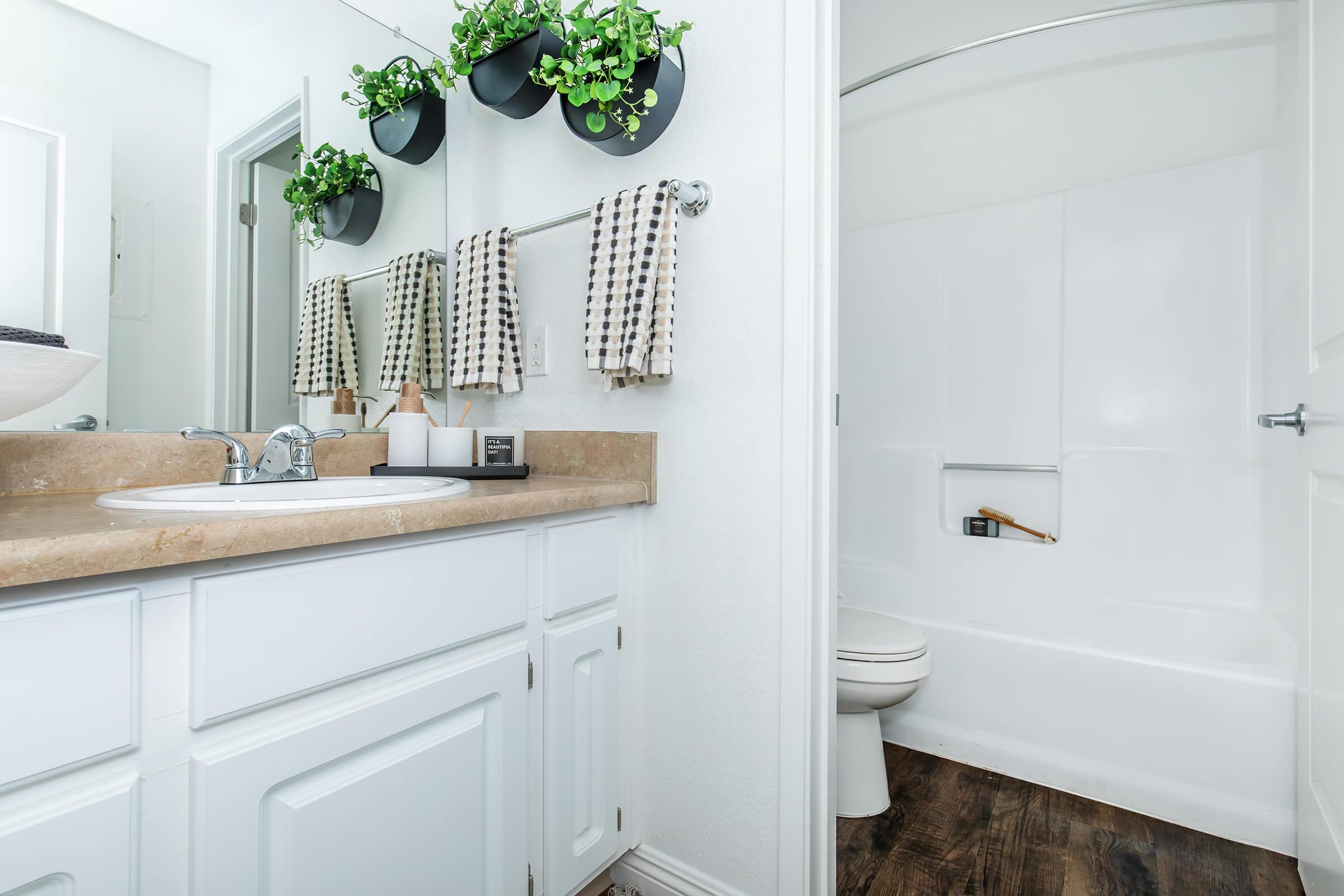 A modern bathroom featuring a beige countertop with a sink, a wall-mounted mirror, and decorative plants in hanging planters. The decor includes patterned towels on a towel rack and a white bathtub with a shower. The floor has dark wood finishes, creating a contemporary and clean aesthetic.