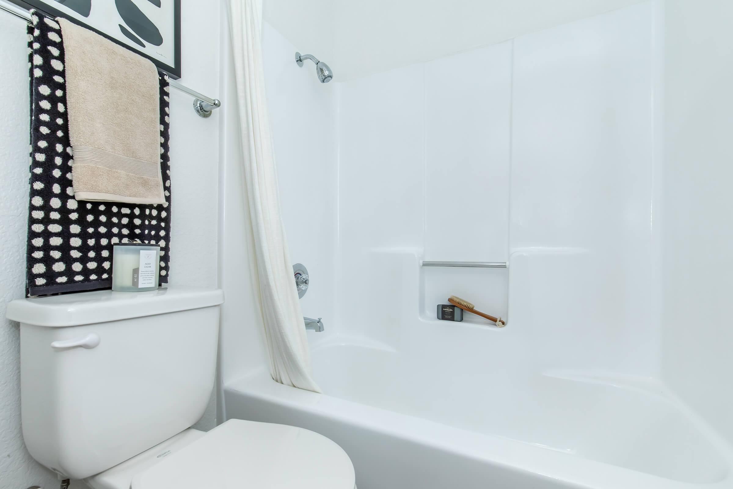 A clean, modern bathroom featuring a white bathtub with a shower curtain. There's a towel hanging on a rack, a candle on the edge of the tub, and a toothbrush holder visible. The walls are plain, creating a bright and airy atmosphere.