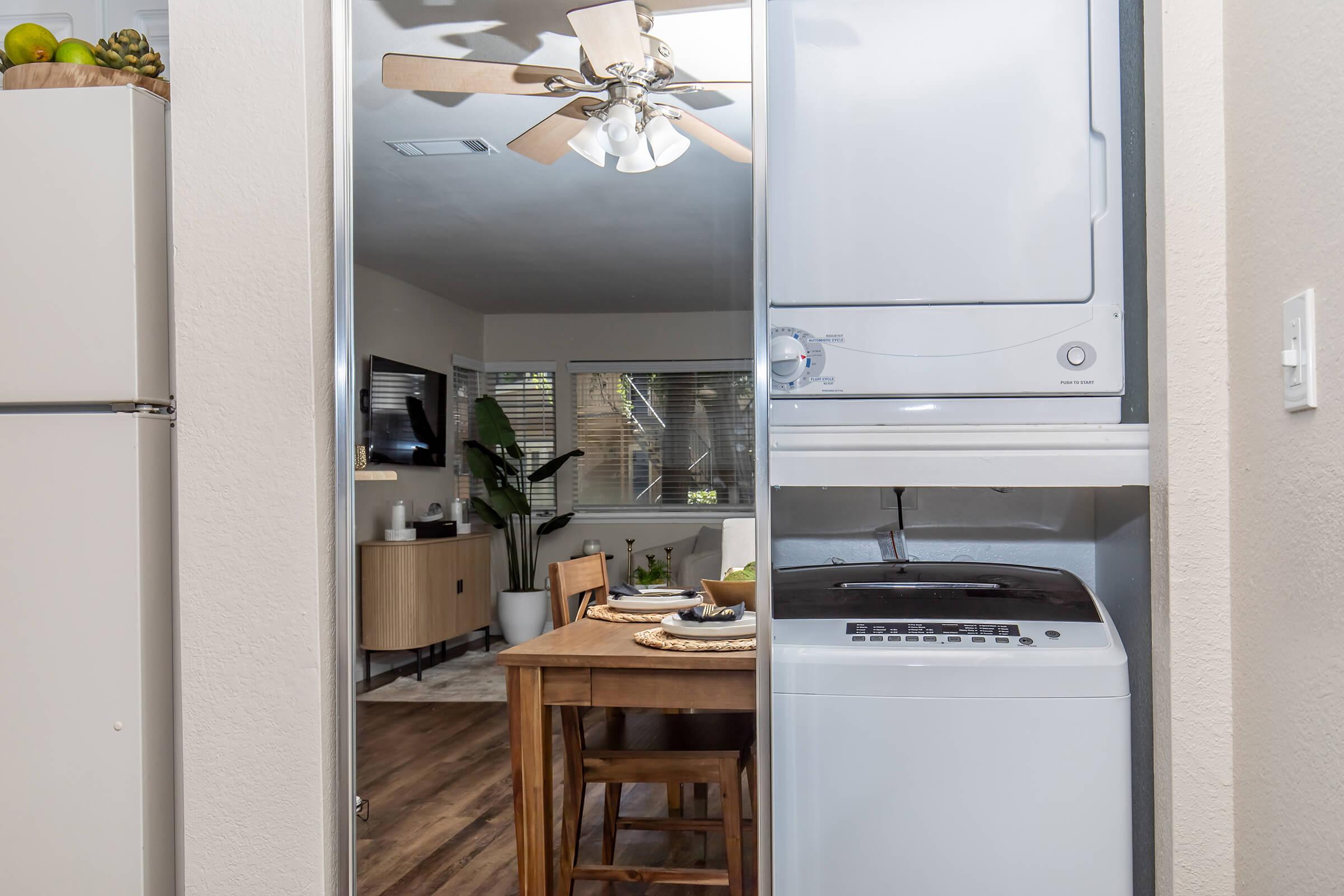 View into a modern kitchen with a dining table and chairs. A stacked washer and dryer are visible on the left. The space features a ceiling fan, a television in the background, and a plant, creating a cozy and functional atmosphere.