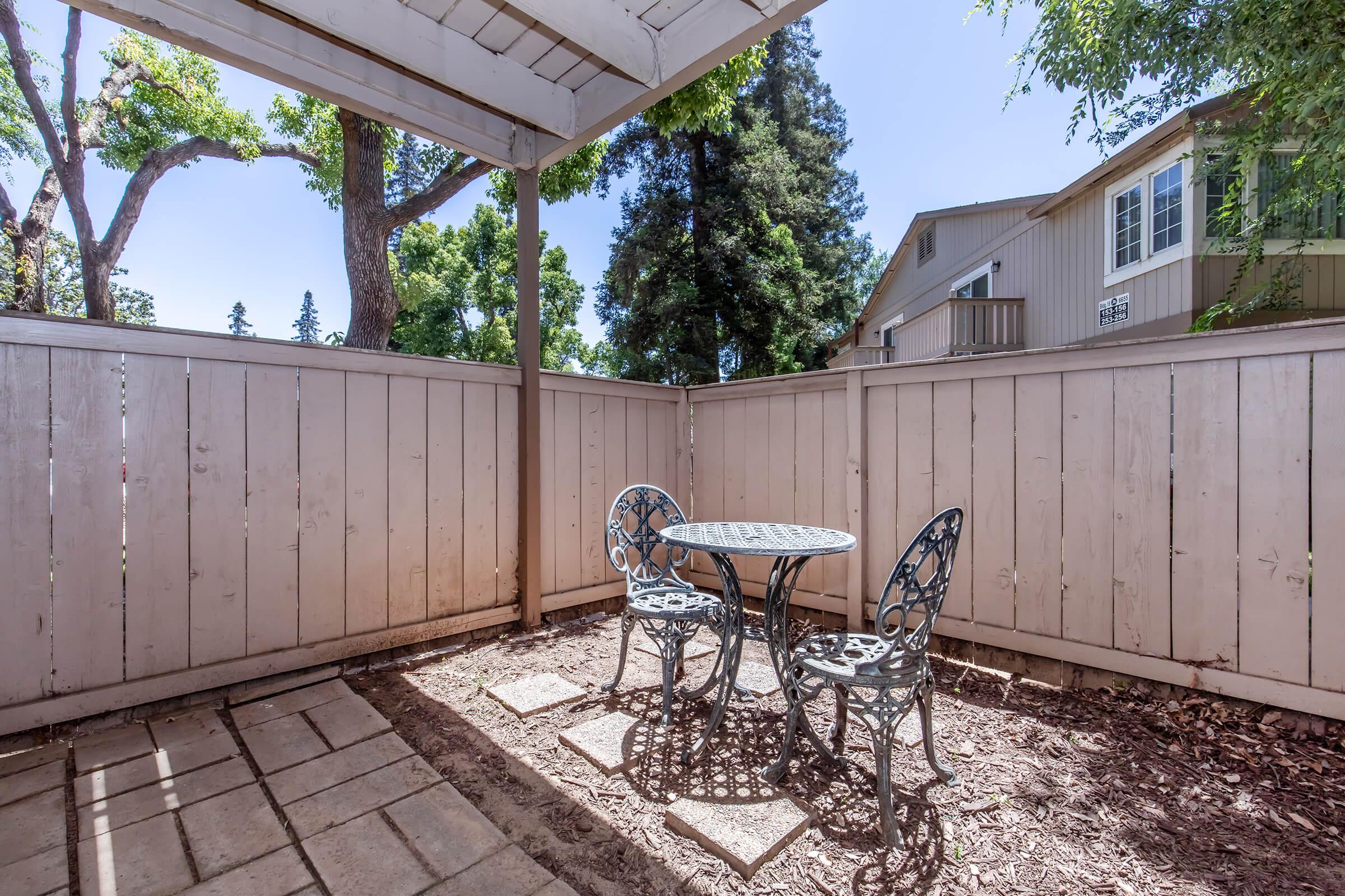 A small outdoor patio area enclosed by wooden fencing, featuring a round metal table with two matching chairs. The space is shaded by a roof and surrounded by trees, creating a tranquil setting for relaxation. Sunlight filters through, illuminating the area.