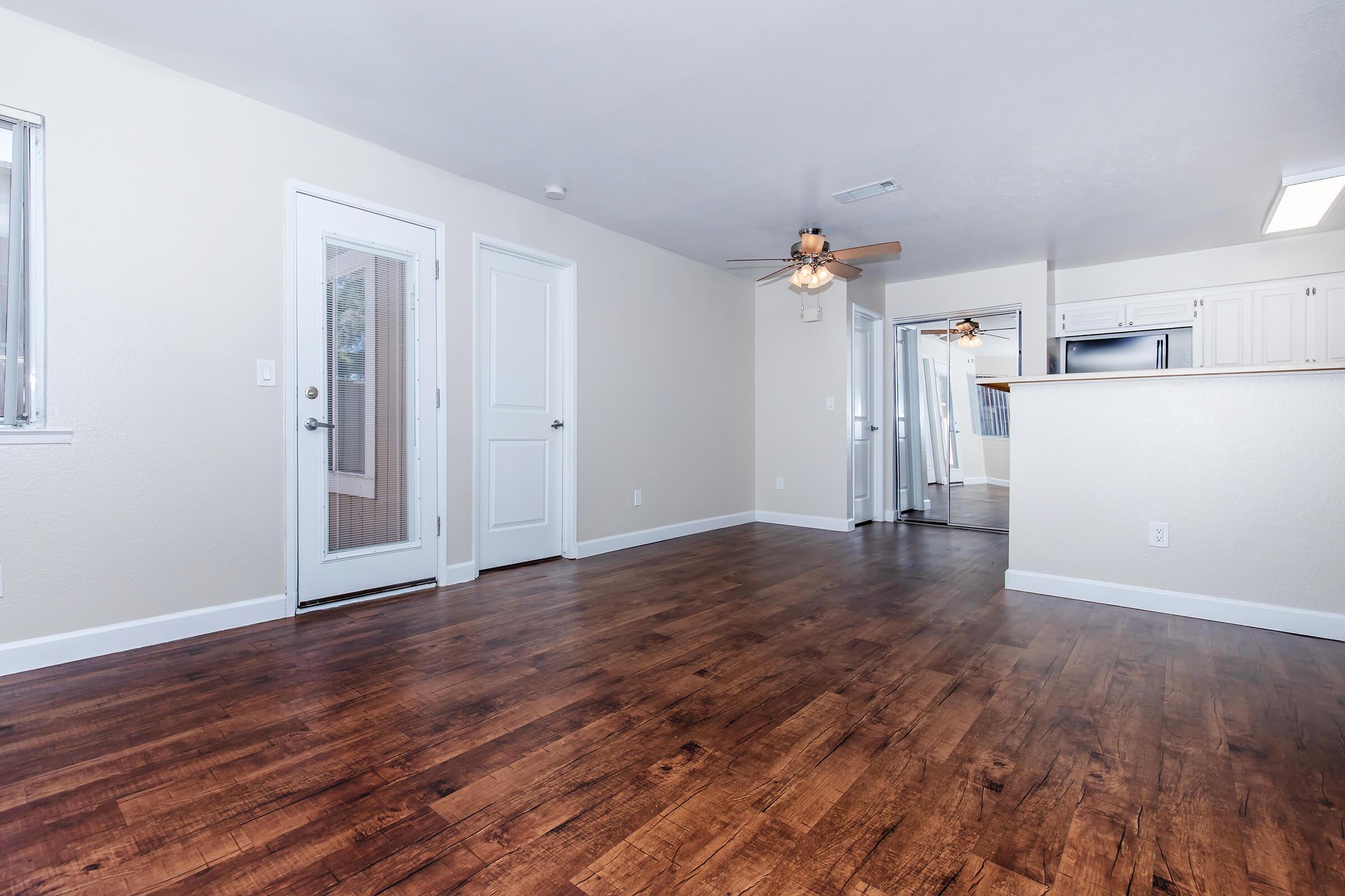 A spacious, well-lit interior of an apartment featuring hardwood flooring, a ceiling fan, and light-colored walls. There are two open doors leading to other rooms, and a kitchen area visible in the background, creating an inviting atmosphere.