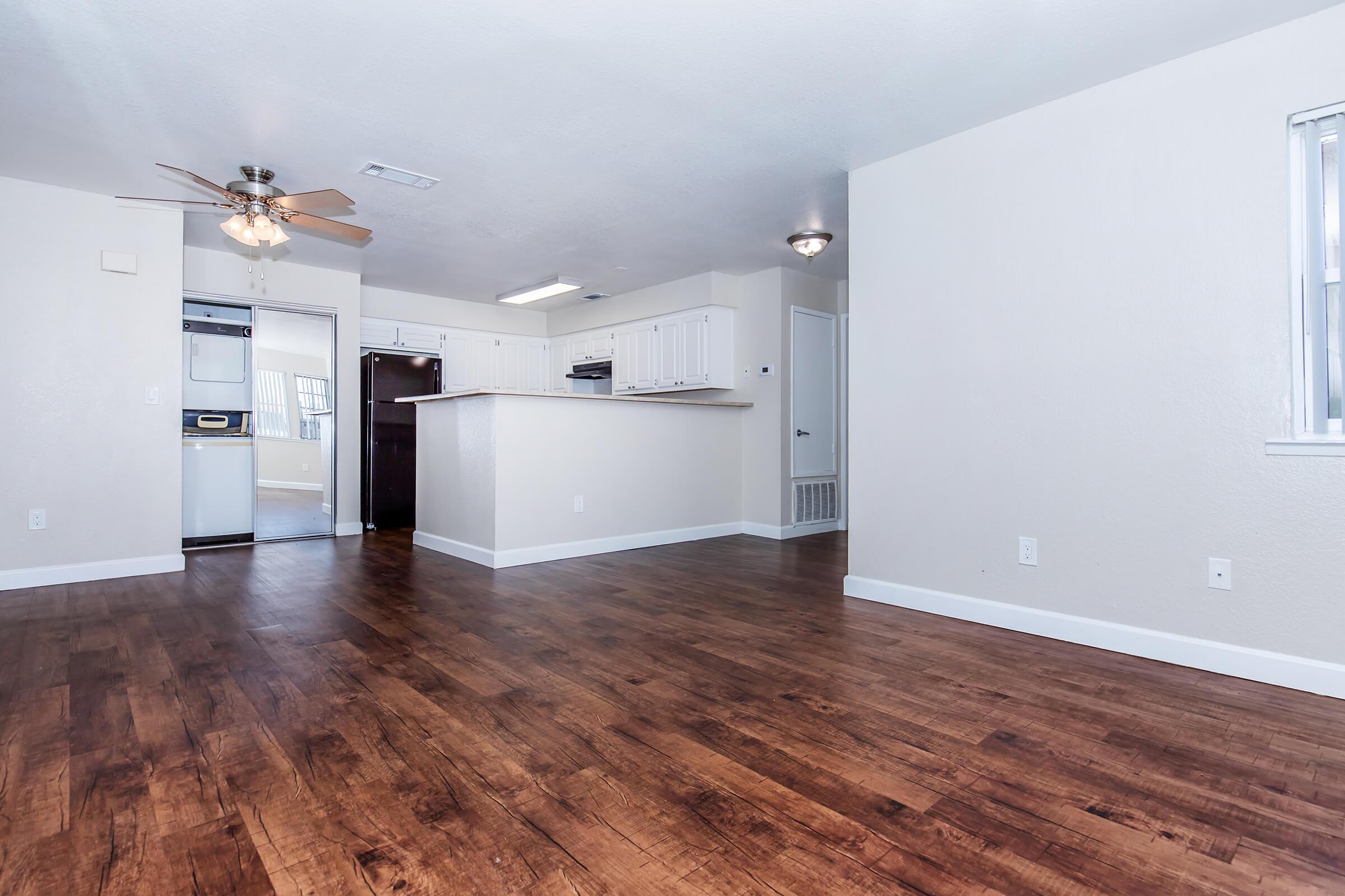 Spacious living area featuring light-colored walls and hardwood flooring. A ceiling fan is visible, and the open layout connects to a kitchen area with white cabinets. Adjacent is a laundry space with appliances. Natural light enters through a window, creating a bright and inviting atmosphere.
