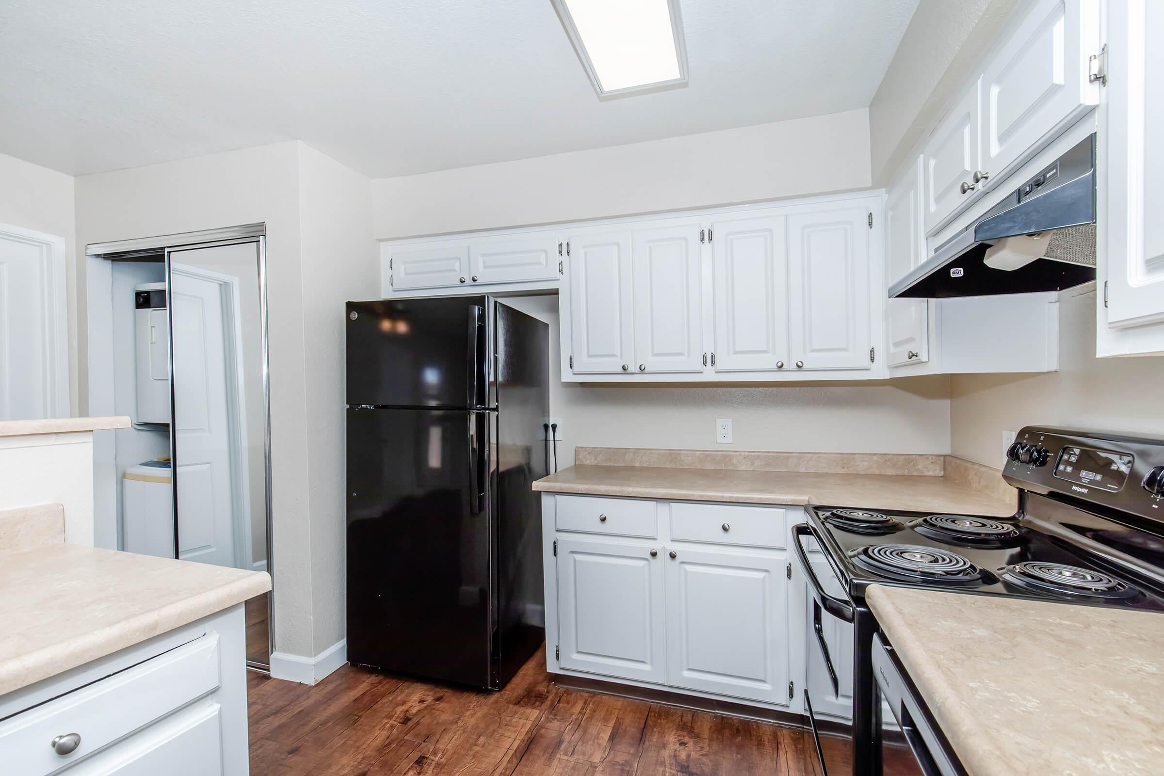A modern kitchen featuring white cabinets, a black refrigerator, a black stove with an exhaust hood, and light-colored countertops. The room has a neutral wall color and wooden flooring, creating a bright and inviting atmosphere. A nearby doorway leads to additional space.