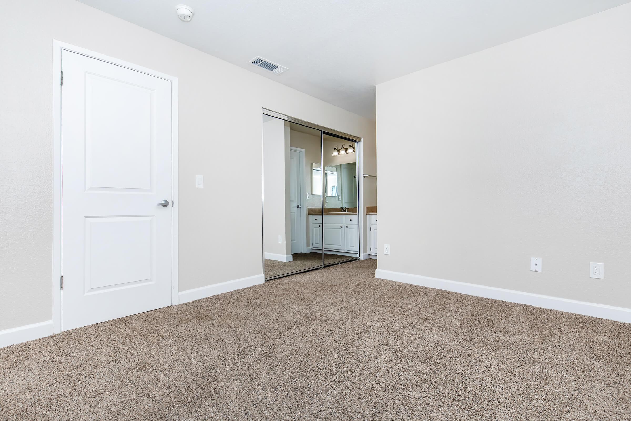A clean, unfurnished room with light beige walls and carpeting. A white door on the left leads outside, and a mirrored closet door is visible on the right. A bathroom area with a sink and vanity is partially visible in the mirror. Natural light brightens the space, creating an inviting atmosphere.