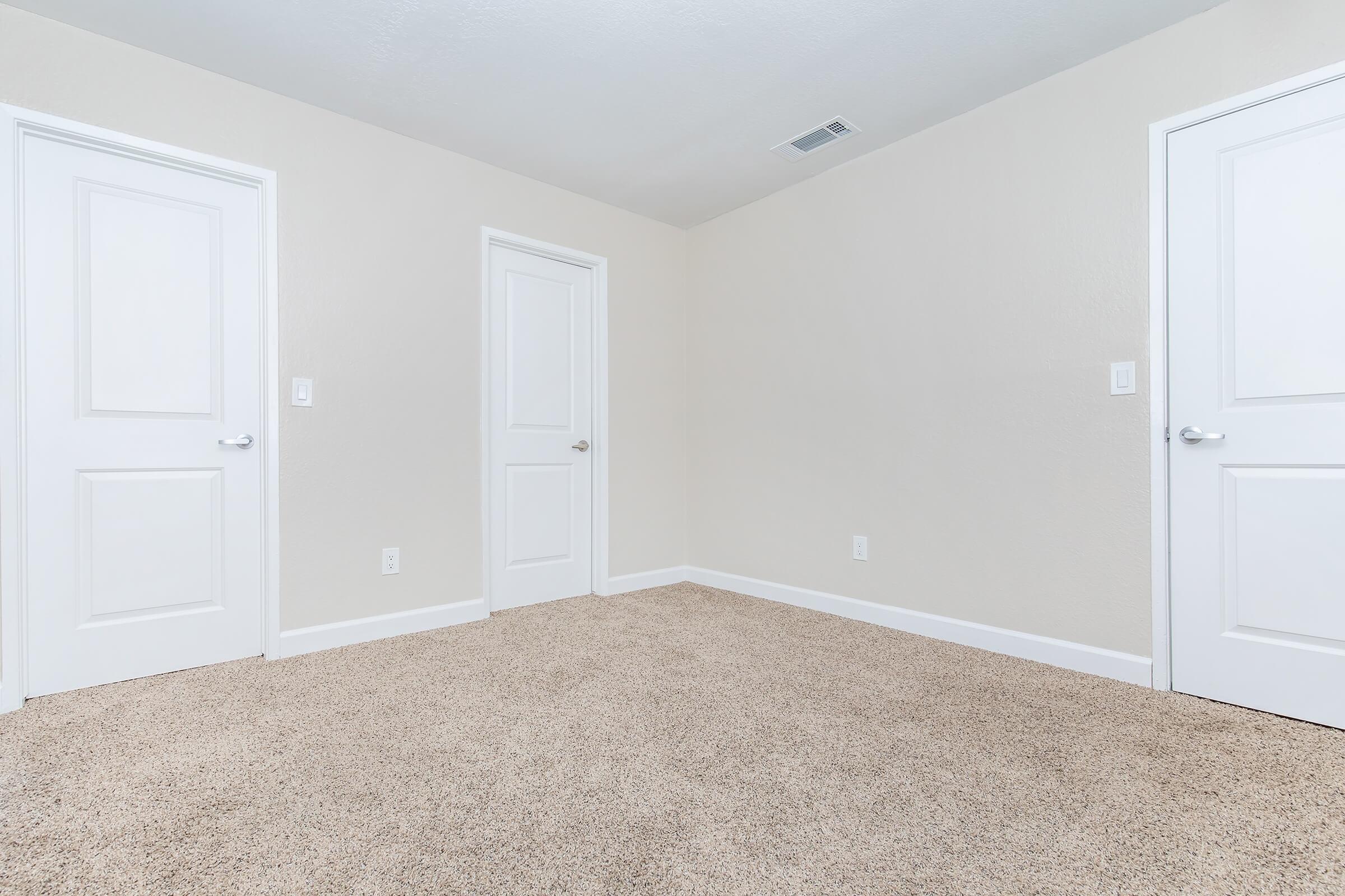 A clean, empty room with light-colored walls and a plush carpet. It features two white doors on opposing walls, with no furniture or decor in the space, creating a bright and open atmosphere. The ceiling is simple and the lighting appears even, emphasizing the room's spaciousness.