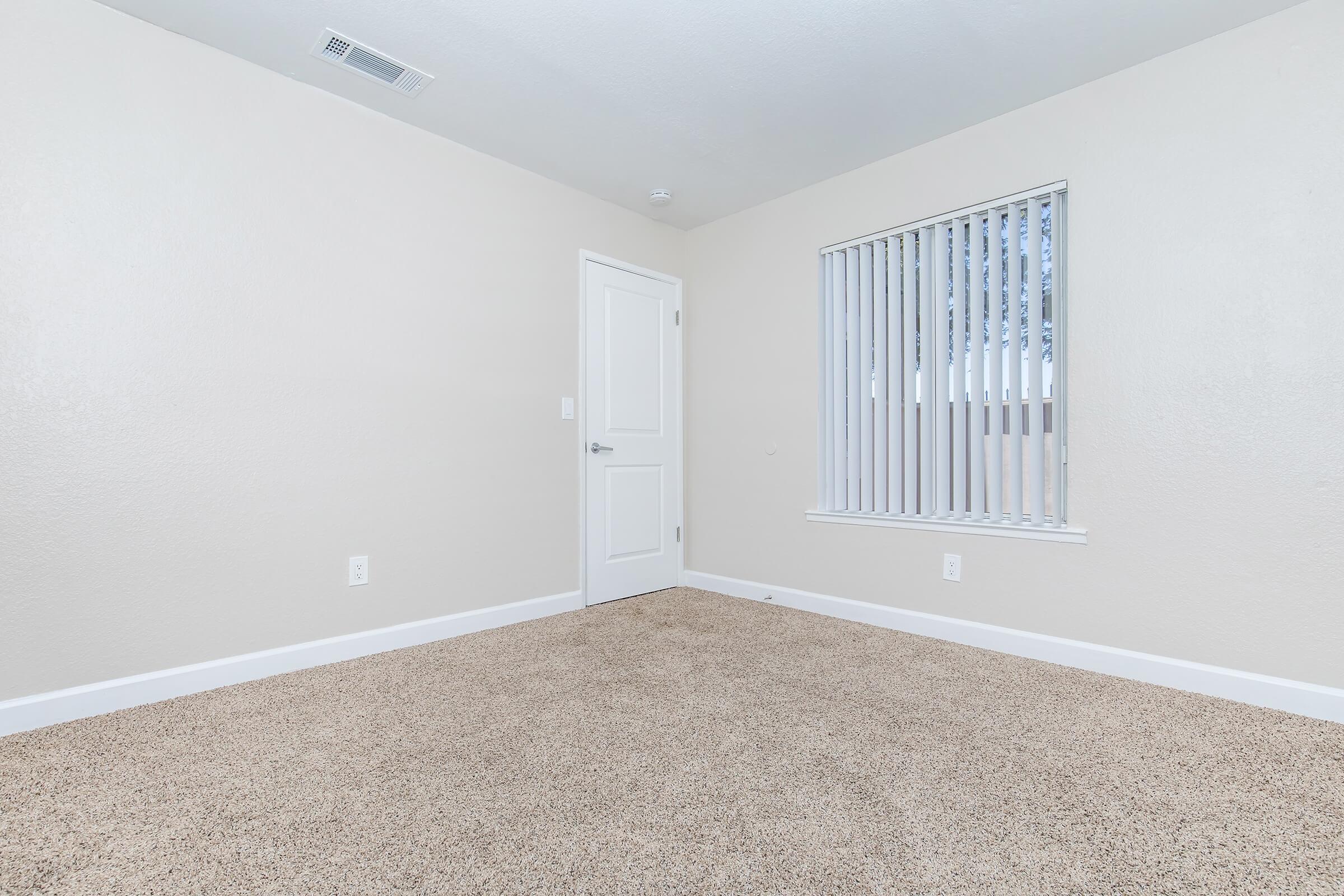 A vacant room with beige carpet, freshly painted light-colored walls, and a single window with white vertical blinds. The door is closed, and there are no furnishings or decorations visible. The overall appearance is clean and minimalistic.