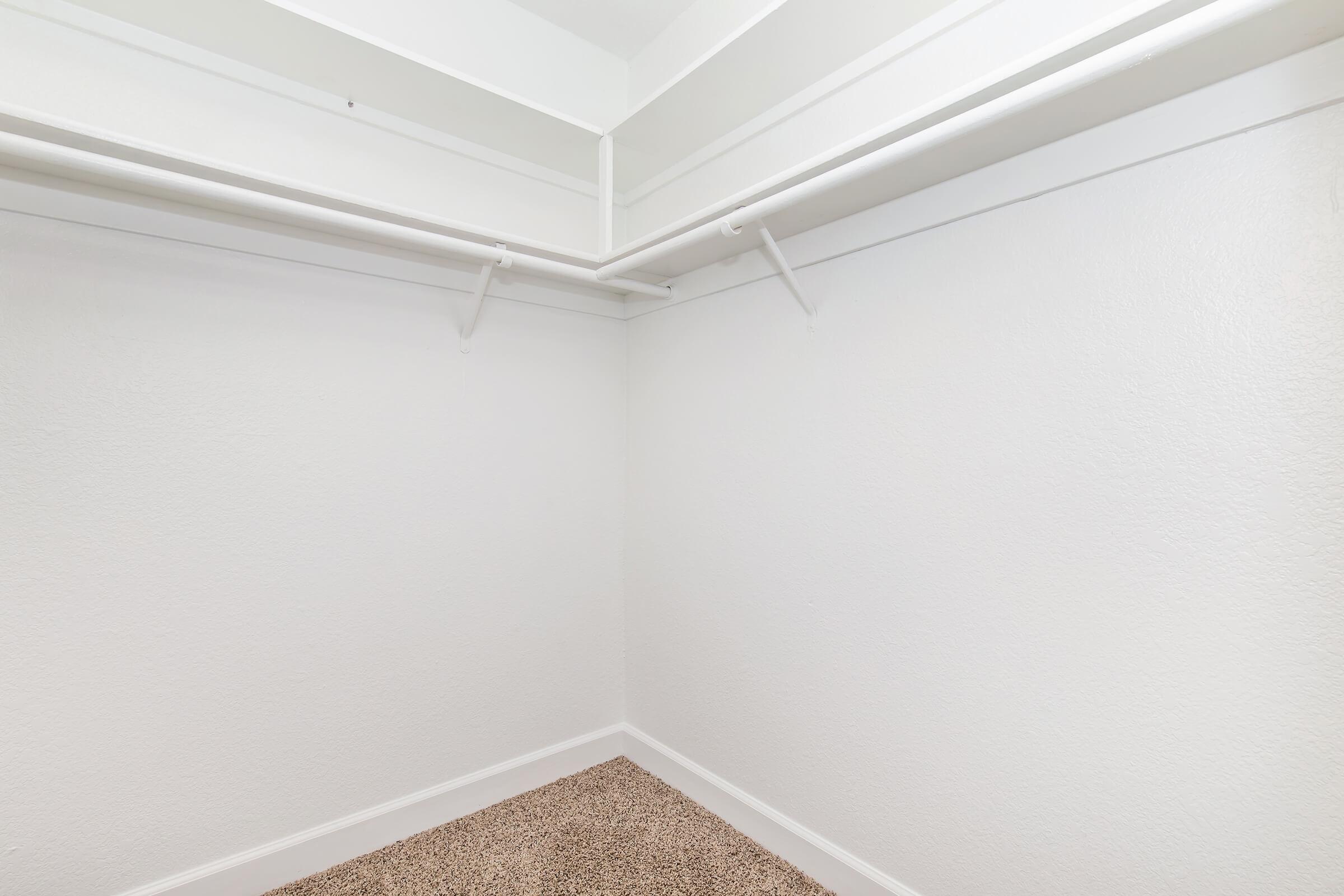 Empty closet with white walls and carpeted floor. Two white hanging rods are installed on the walls, providing space for clothing or storage. The overall look is clean and minimalist, with no items or decorations present.