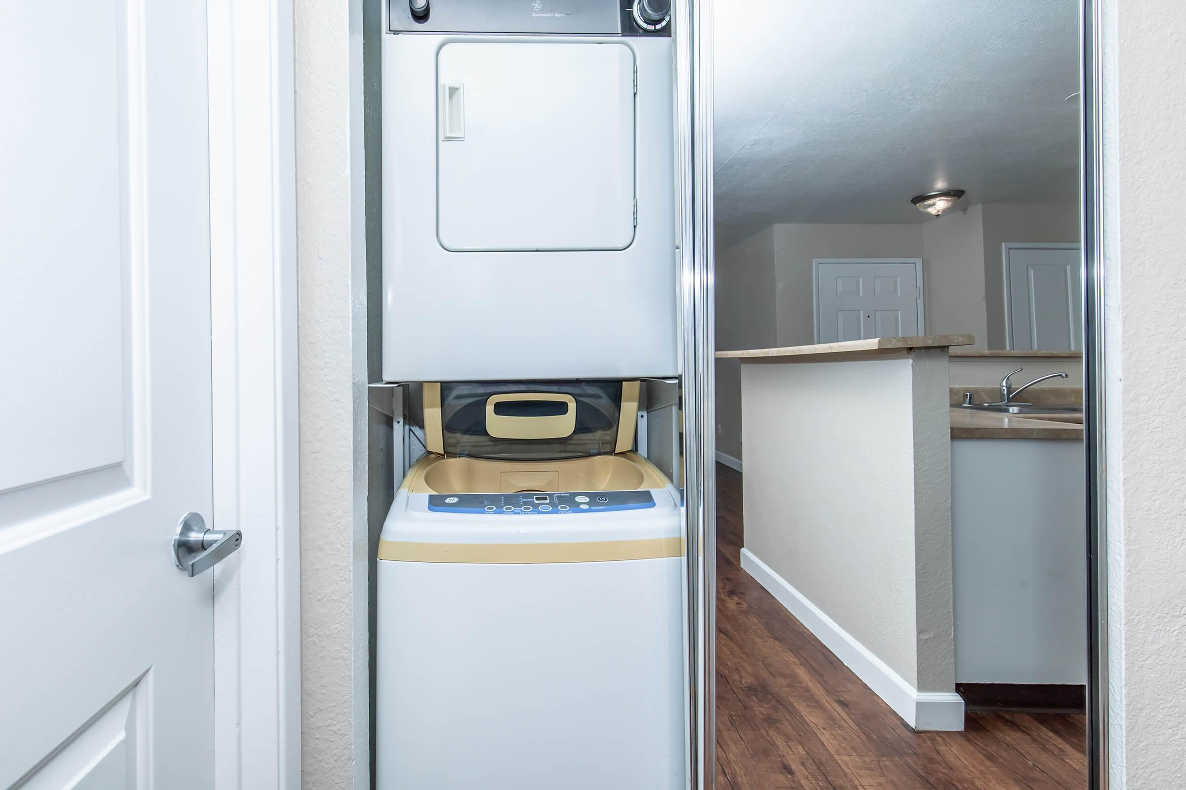 A stacked washer and dryer unit in a hallway, with a view into an adjacent kitchen area featuring a countertop and sink. The walls are light-colored, and the floor is brown wood. The doorway is partially visible on the left.