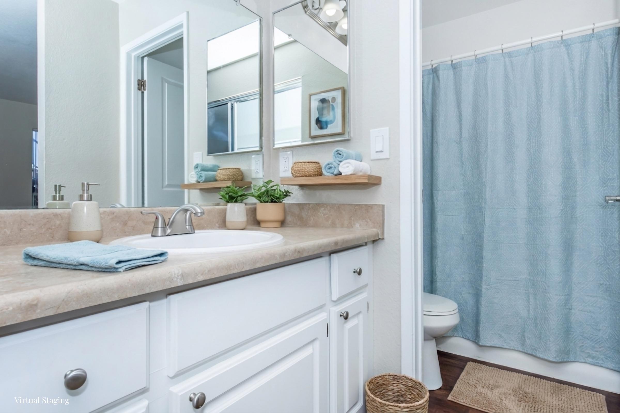 A bright and clean bathroom featuring a light-colored countertop with a sink, neatly arranged toiletries, and a small potted plant. There are mirrors above the sink, and a light blue shower curtain hangs in front of a shower. The floor has a woven mat, enhancing the cozy feel of the space.