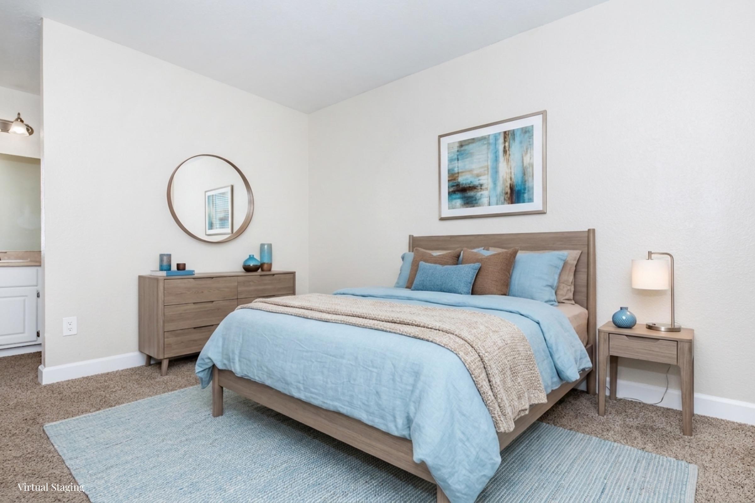 A serene bedroom featuring a light wooden bed with blue and beige bedding, a round mirror above the nightstand, and a dresser. The walls are painted in a soft color, and decorative items in various shades of blue are placed throughout the room, creating a calming atmosphere.