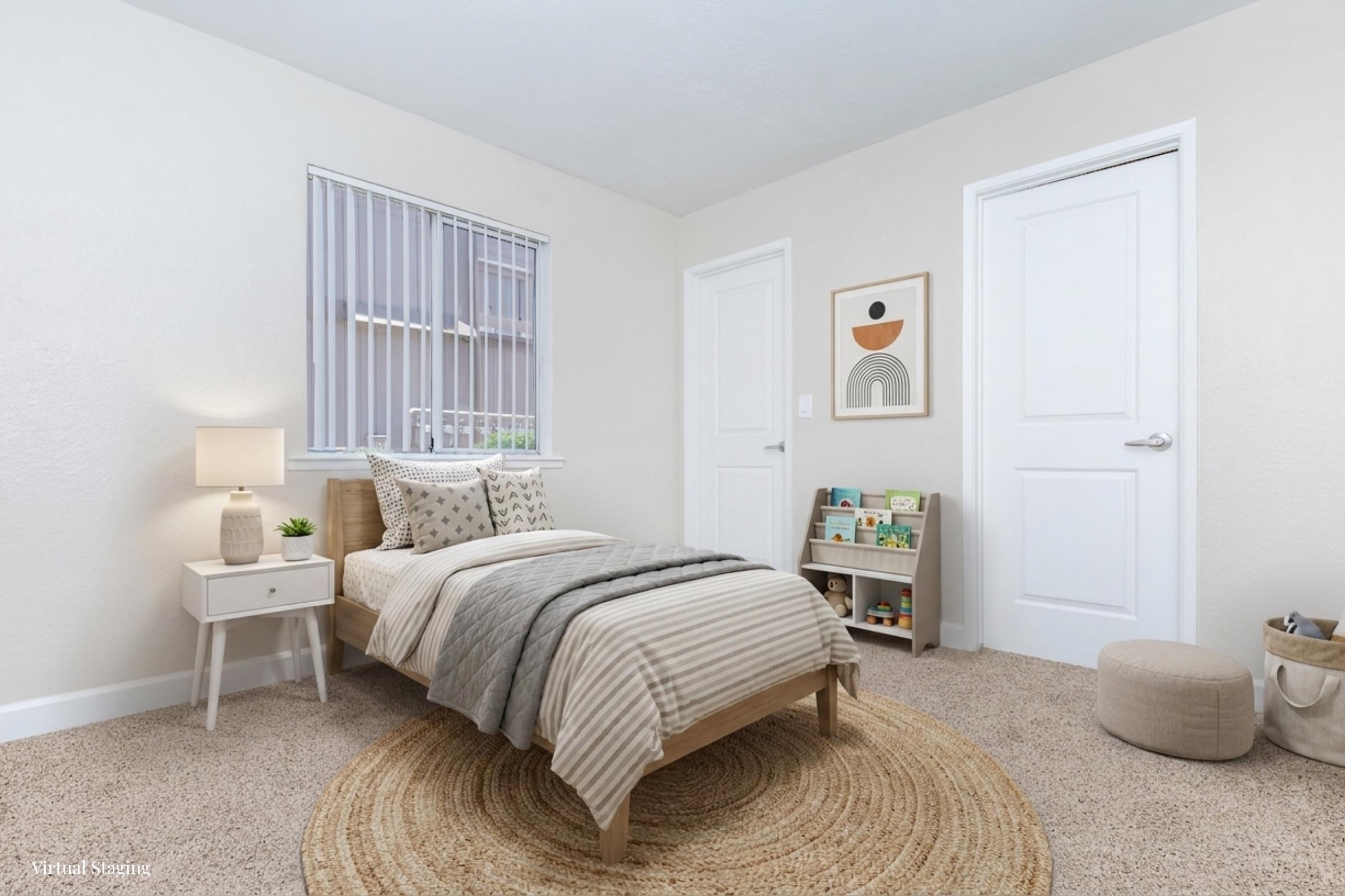 A small, well-lit bedroom featuring a twin bed with beige and gray bedding, a nightstand with a lamp, and a round jute rug. There is a window with blinds, a wall art piece, a closed door, and a shelf with books and toys. The carpeted floor adds warmth to the space.