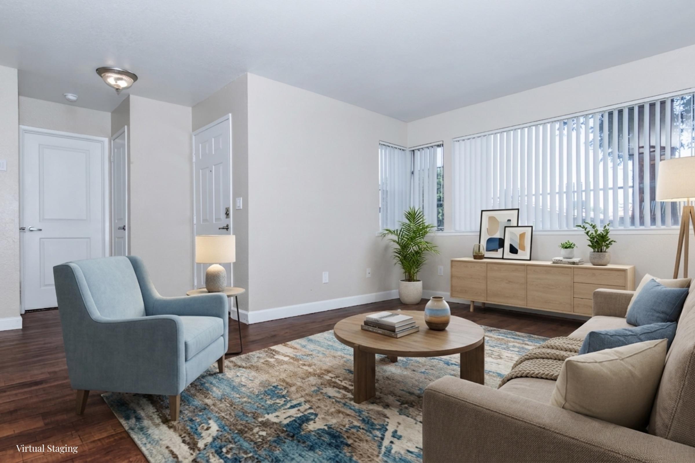A cozy living room featuring a light blue armchair, a beige sofa with decorative pillows, and a round wooden coffee table. The space has a modern design with a patterned area rug, a small plant, and a side table with a lamp. Large windows allow natural light to fill the room, enhancing the welcoming atmosphere.