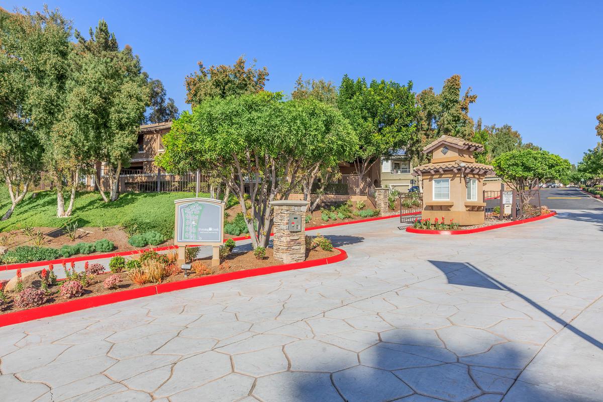 Welcoming entrance to a residential community featuring a landscaped driveway with red-painted edges, lush greenery, and a stone-styled kiosk or entry point. The clear blue sky above enhances the bright and inviting atmosphere of the area.