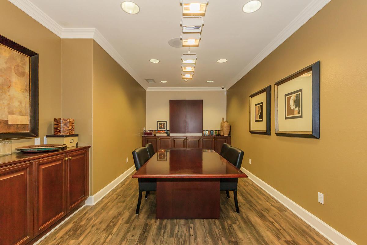 A modern conference room with a dark wood table and black chairs. The walls are painted a warm beige, accented with framed artwork. Track lighting provides bright illumination. A cabinet and shelving unit are visible in the background, featuring decorative items. The overall atmosphere is professional and inviting.