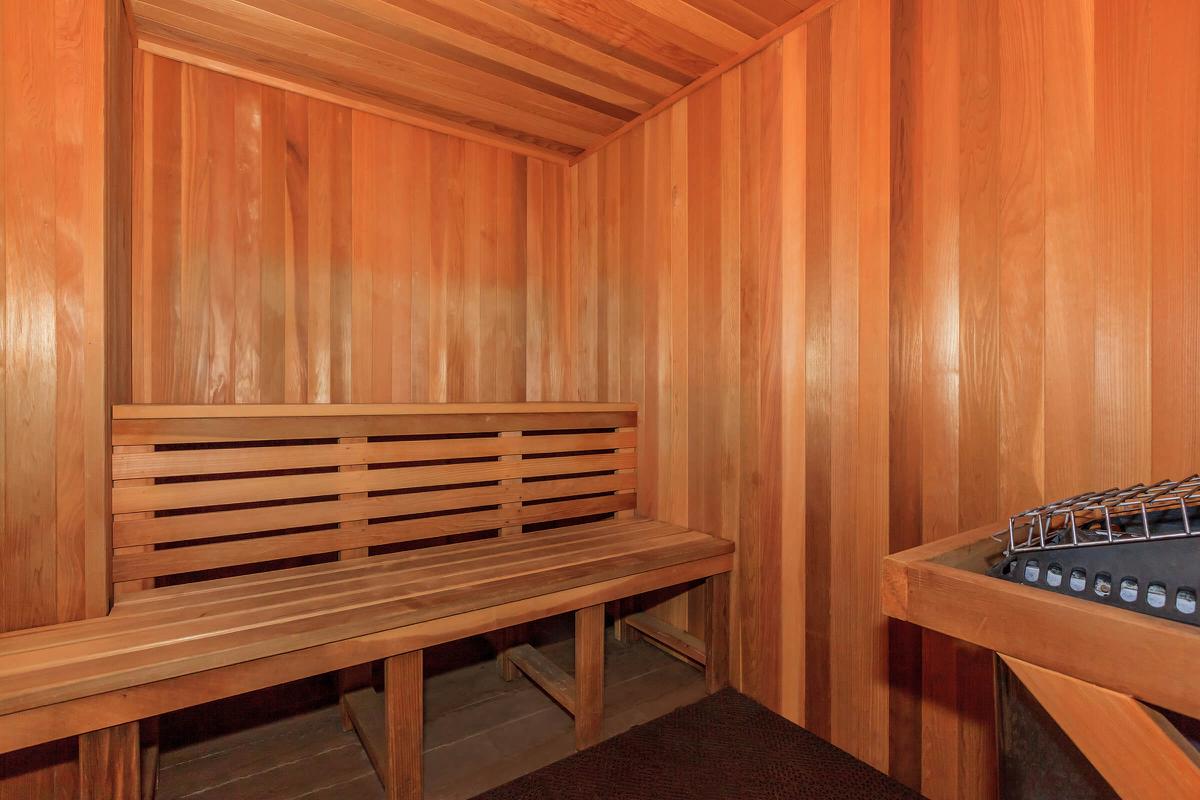 Interior of a wooden sauna featuring bench seating along the walls, warm lighting, and a stone heater in the corner. The walls and ceiling are lined with smooth, light-colored wood, creating a cozy and relaxing atmosphere.