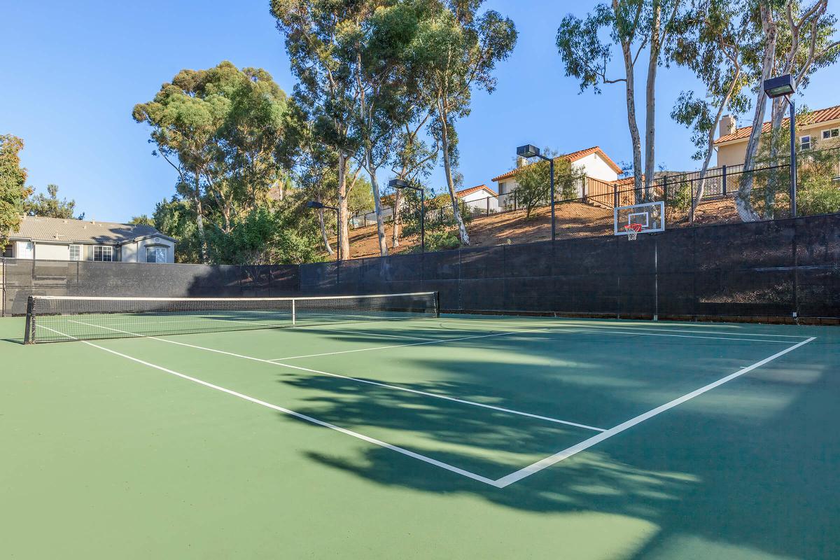 A vacant tennis court surrounded by trees, with a clear sky overhead. The court features a net in the center, painted in green, and has white lines marking the boundaries. In the background, there are residential buildings and a basketball hoop attached to a fence. The scene conveys a tranquil outdoor sports area.