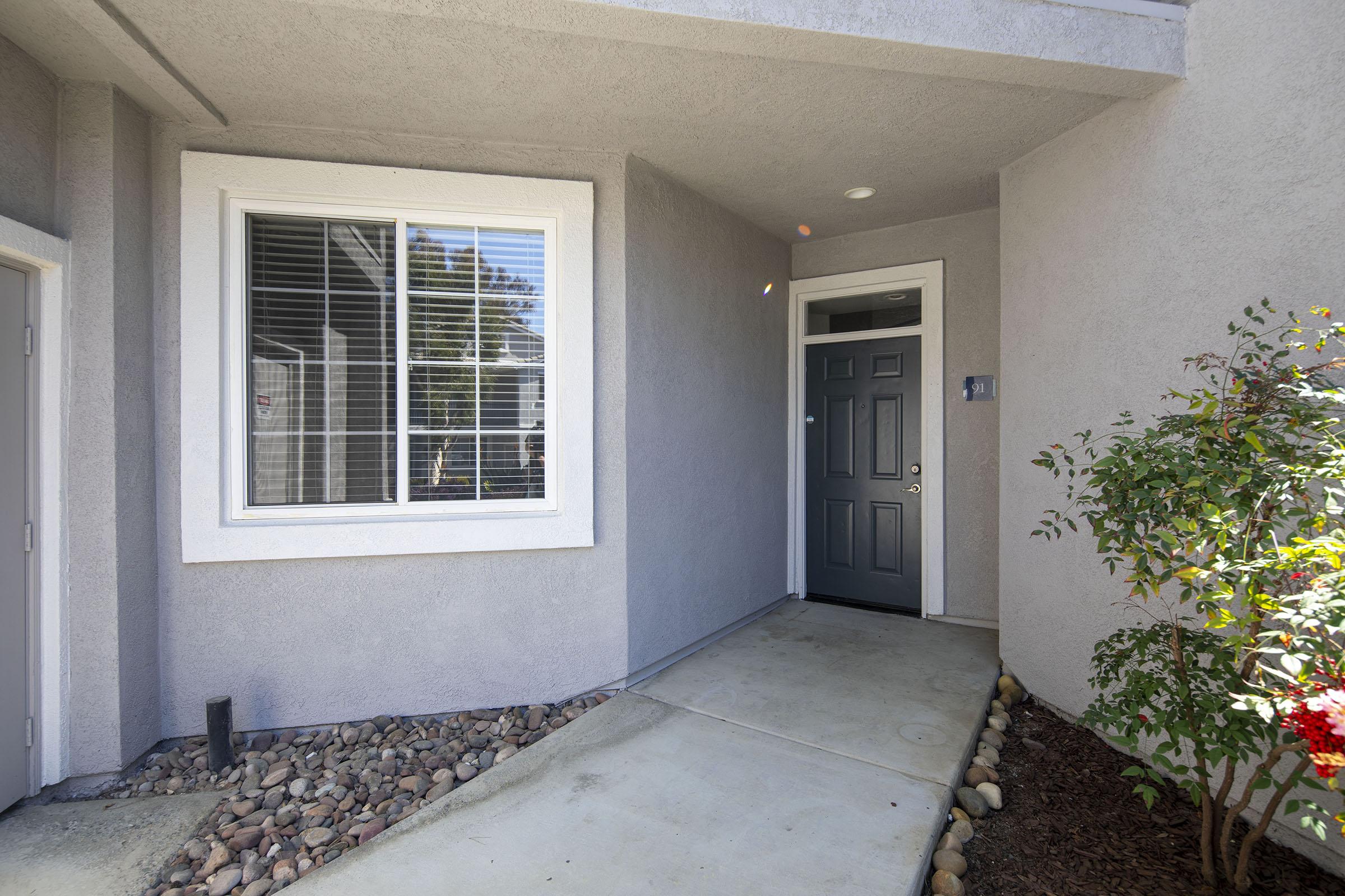 Entrance to an apartment featuring a gray exterior wall, a dark green front door, and a well-maintained walkway. There is a window with white trim beside the door, allowing natural light in. The area is landscaped with small rocks and foliage, creating a welcoming atmosphere.