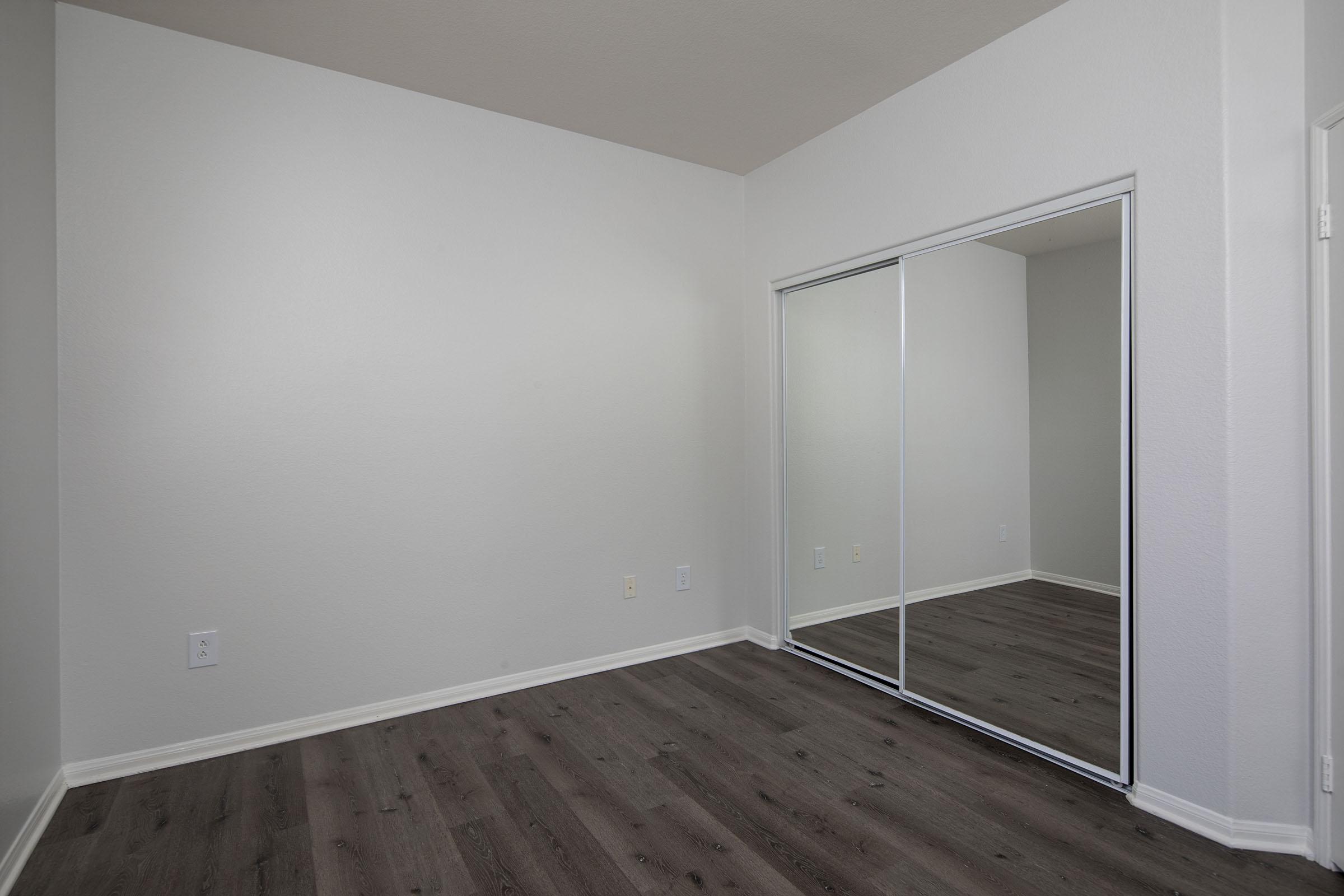 A minimalistic, empty room with light gray walls and a large mirrored closet door. The floor is dark wood laminate. The space is bright and uncluttered, featuring a corner for potential furniture placement.