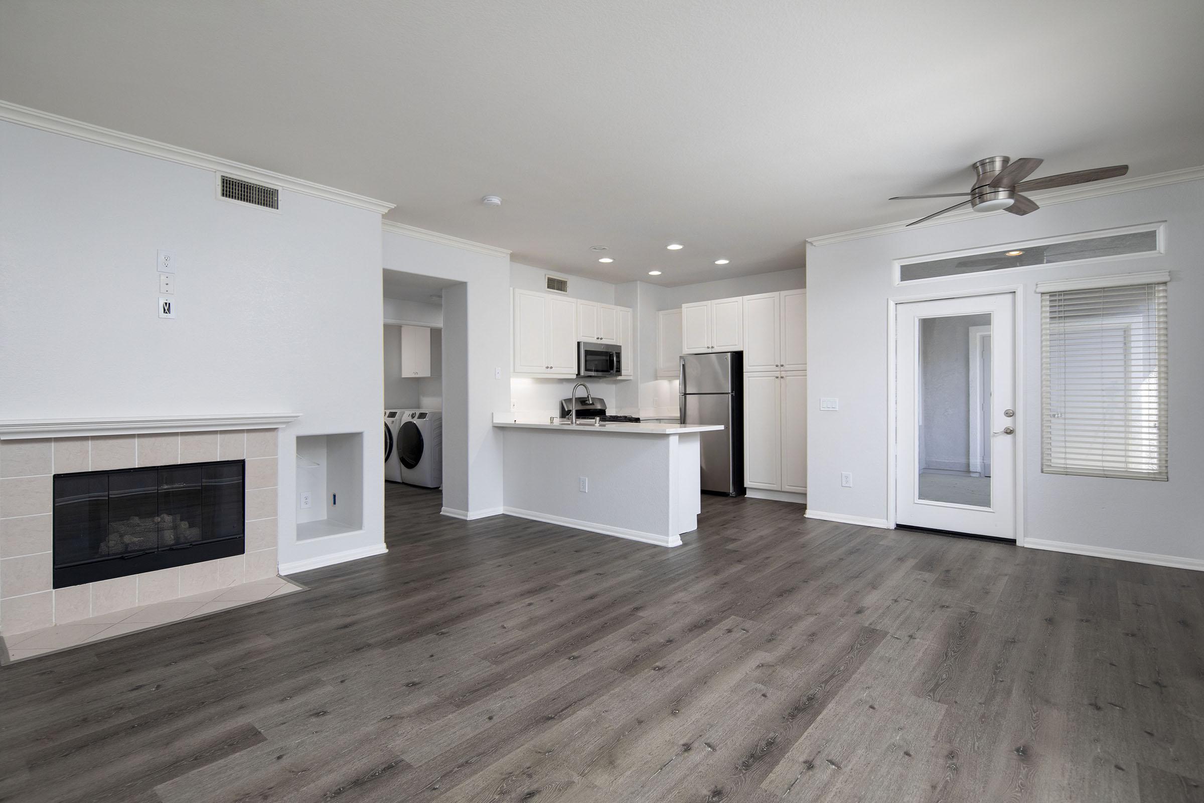 A modern, open-concept living area featuring a light-colored wall, hardwood flooring, a fireplace, and a kitchen with stainless steel appliances. A door leads to an exterior space, and laundry facilities are visible in a separate room. Natural light enters through a window with blinds.