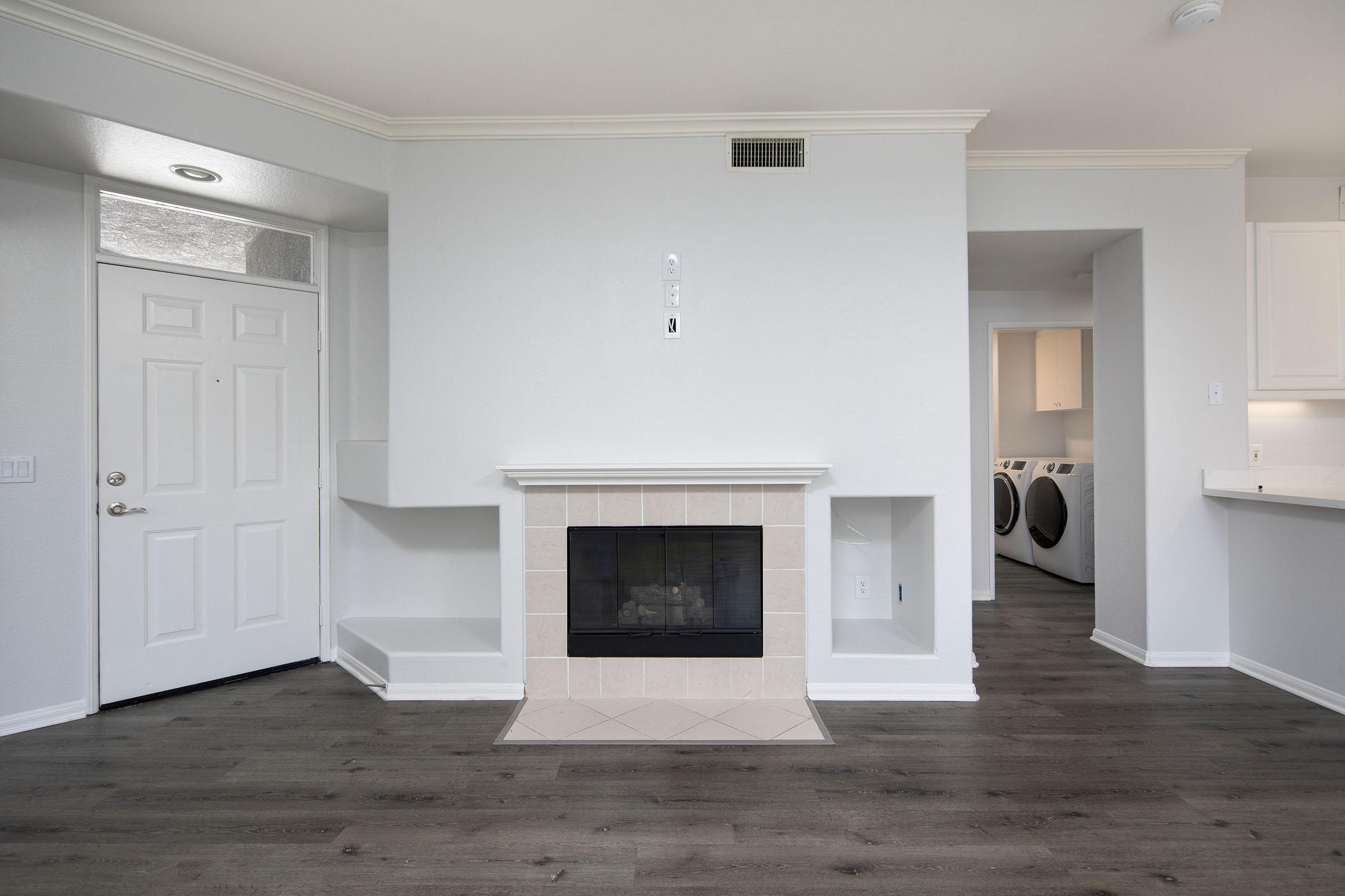 A modern living room featuring a white wall with a fireplace at the center, a door to the left, a small shelf beside the fireplace, and an open view into a laundry area with appliances. The flooring is dark wood, and the space is well-lit with neutral colors throughout.