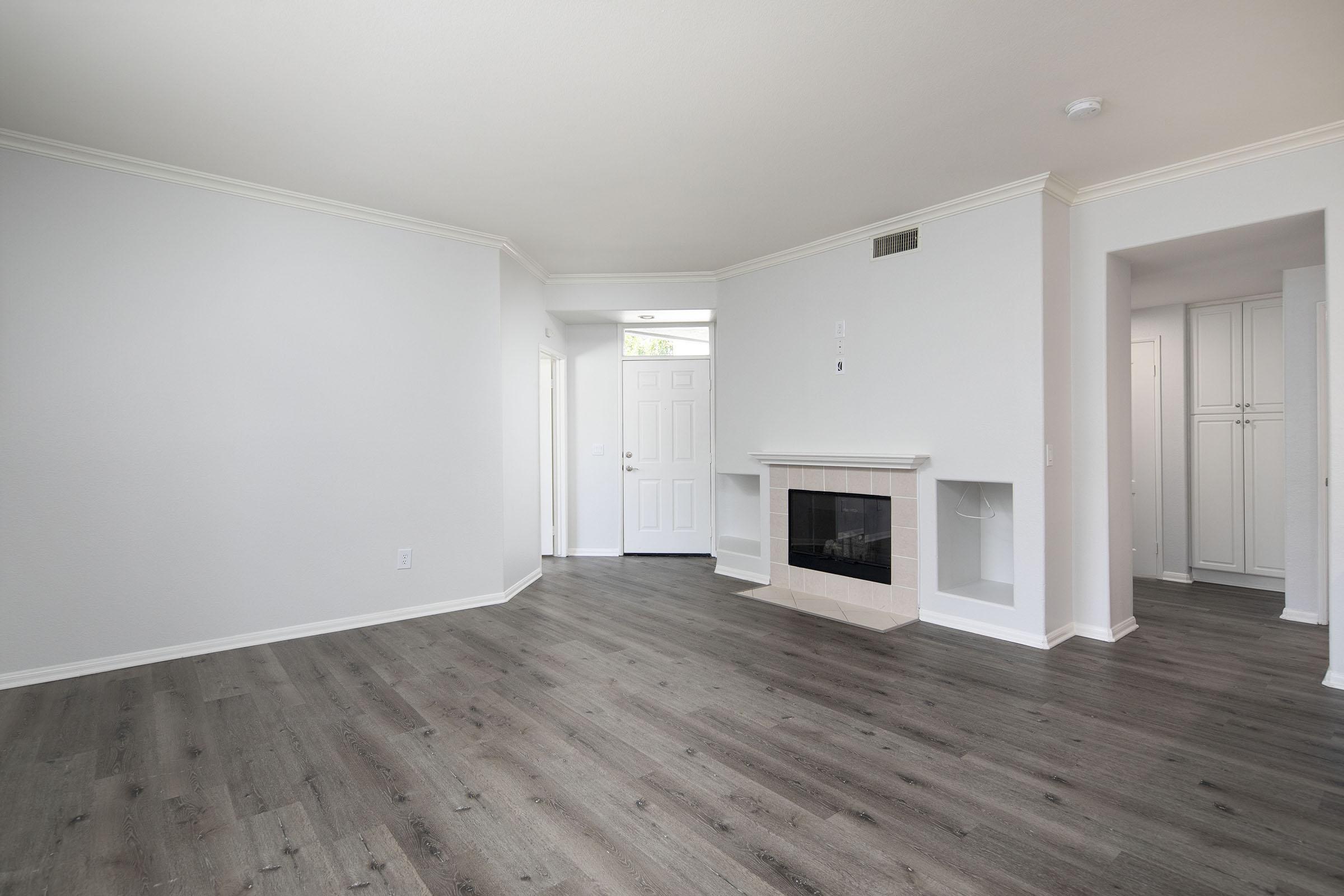 A spacious, empty living room with light gray walls and wooden flooring. There is a white door leading outside, a fireplace with a simple mantle, and a built-in shelf. Adjacent to the living room, there are two doorways leading to other rooms, creating an open layout. Natural light enters from the door.