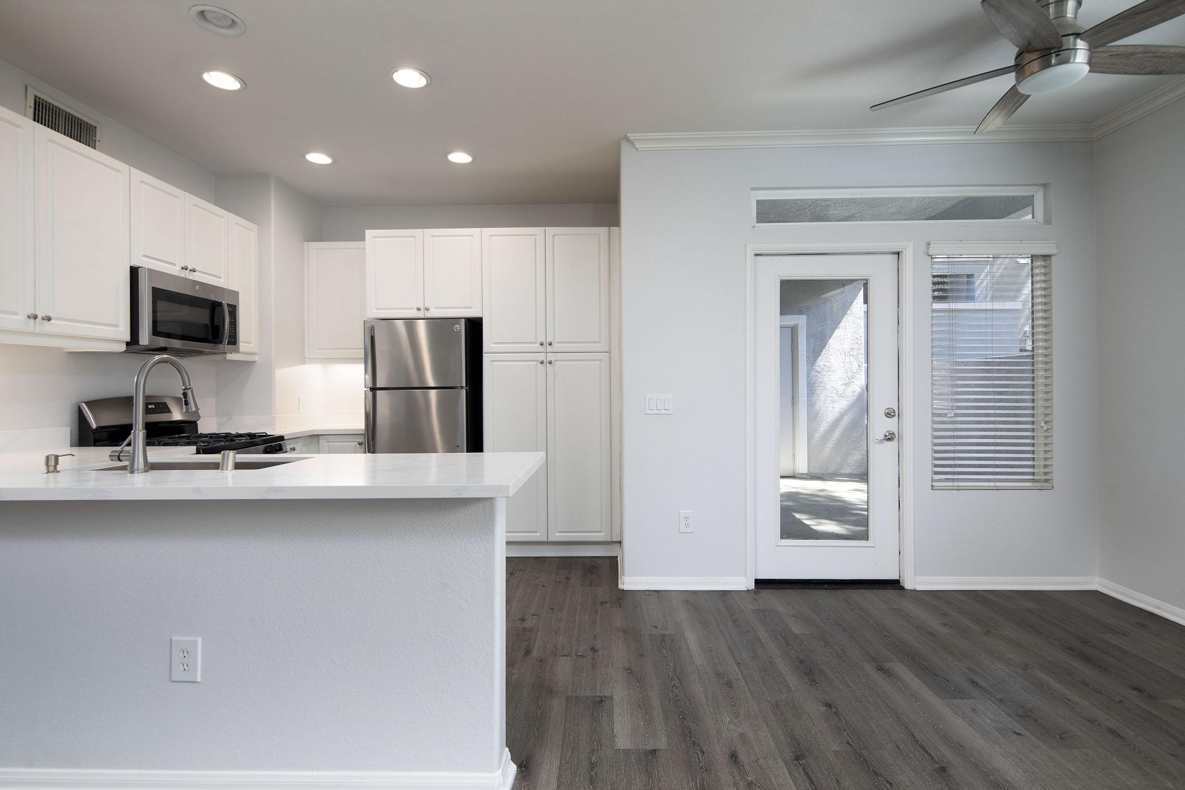 A modern kitchen featuring white cabinetry, stainless steel appliances, and a spacious island. The adjacent living area has a door leading outside, and large windows providing natural light. The flooring is wood-like, creating a cohesive and inviting atmosphere.