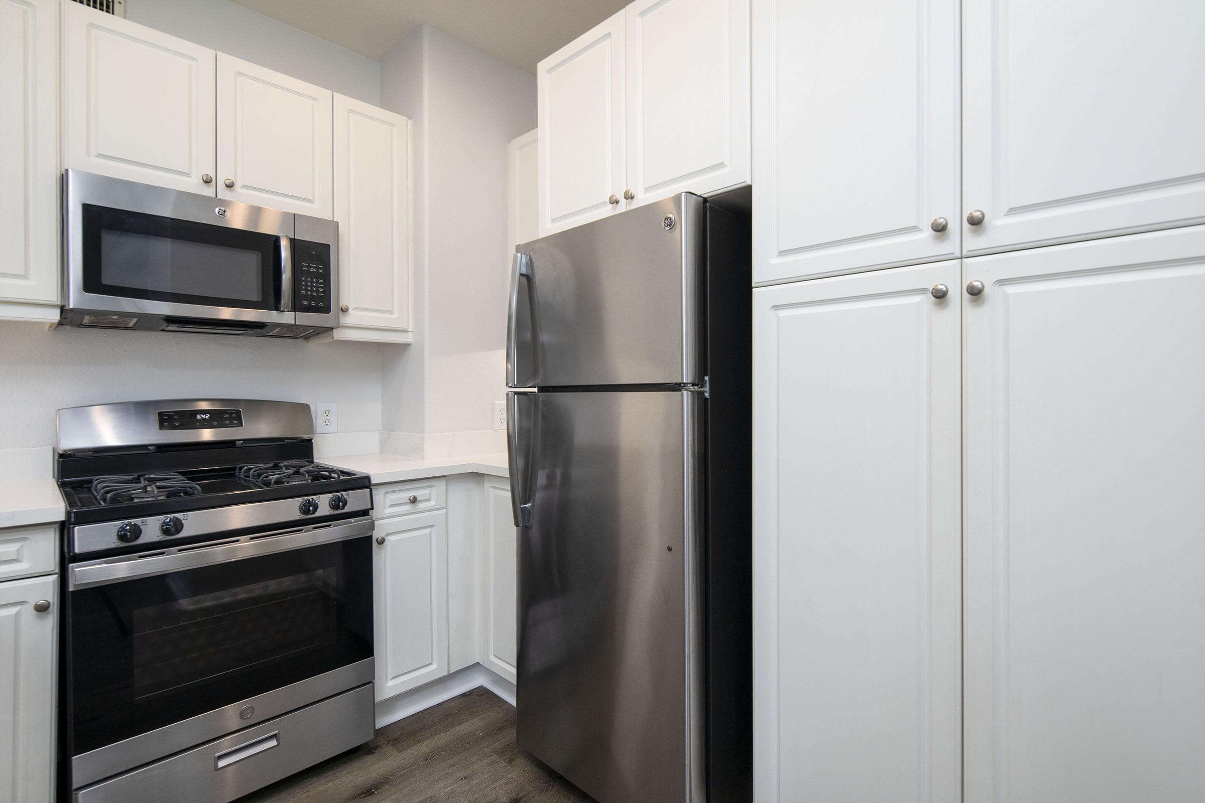 Modern kitchen featuring stainless steel appliances, including a gas stove, microwave, and a refrigerator. The cabinets are white with a clean, minimalist design, complemented by a light-colored countertop. The flooring is dark, adding contrast to the overall bright atmosphere of the space.