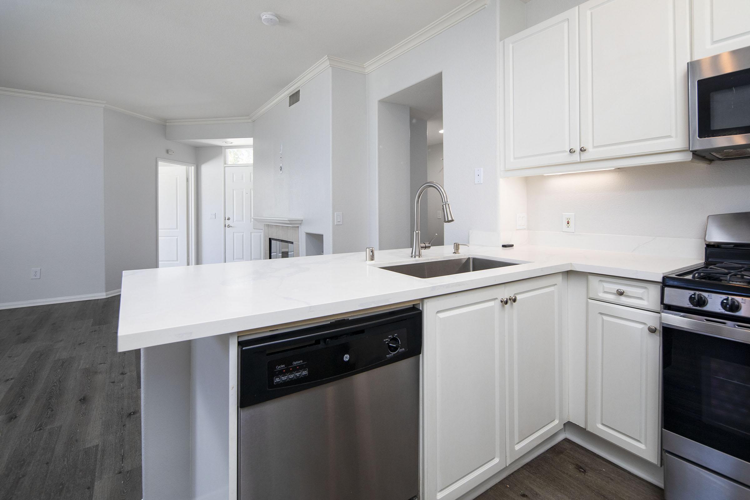 A modern kitchen featuring white cabinetry, a stainless steel dishwasher, and a double sink on a light marble countertop. The background shows an open layout with a view into a living area and a door leading outside, all set in a neutral-toned space with hardwood flooring.