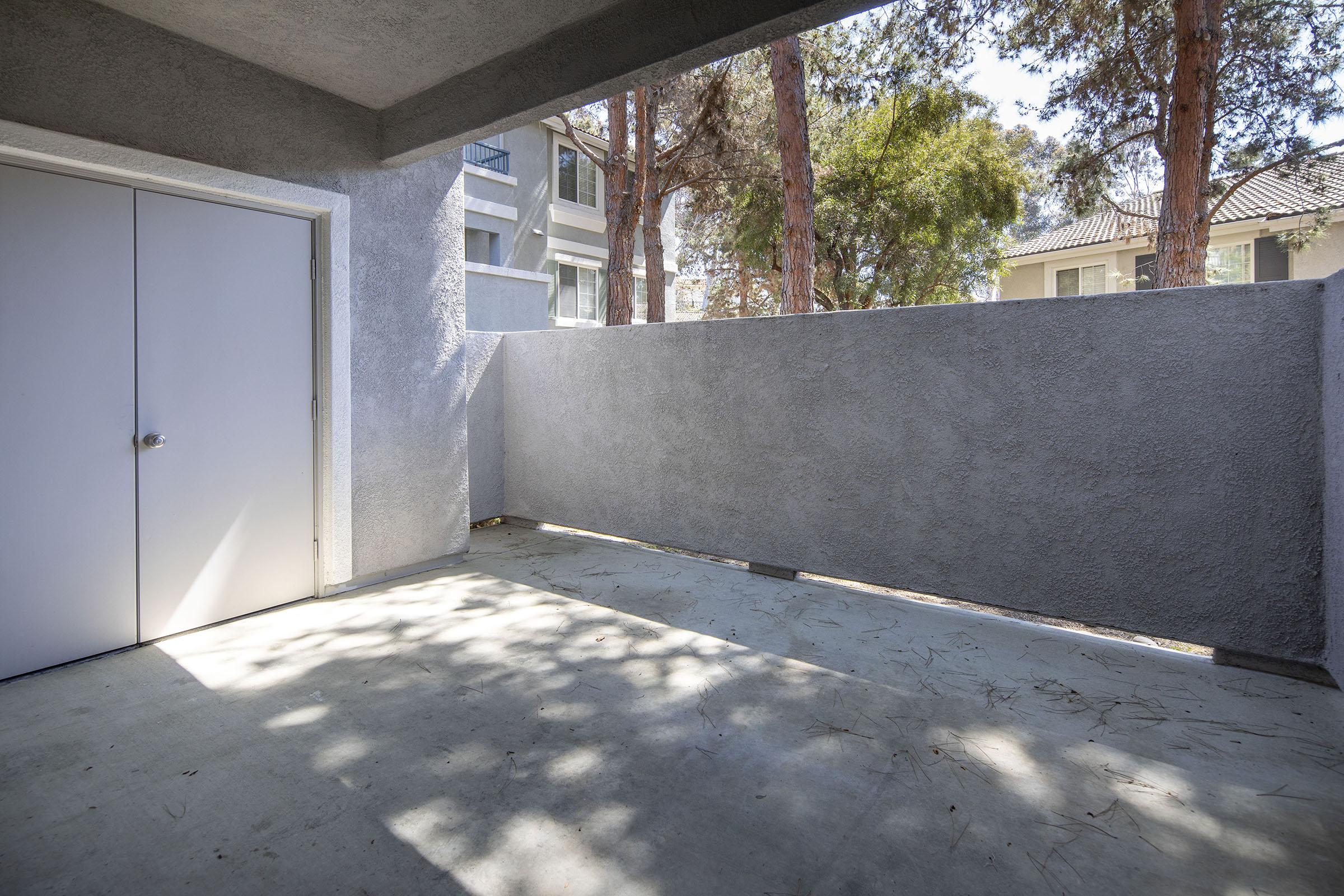 A small, private outdoor patio area with a concrete floor and grey walls. There is a closed white door on the left side, and trees can be seen in the background, providing some shade. Sunlight casts shadows on the floor, creating a tranquil and inviting atmosphere.