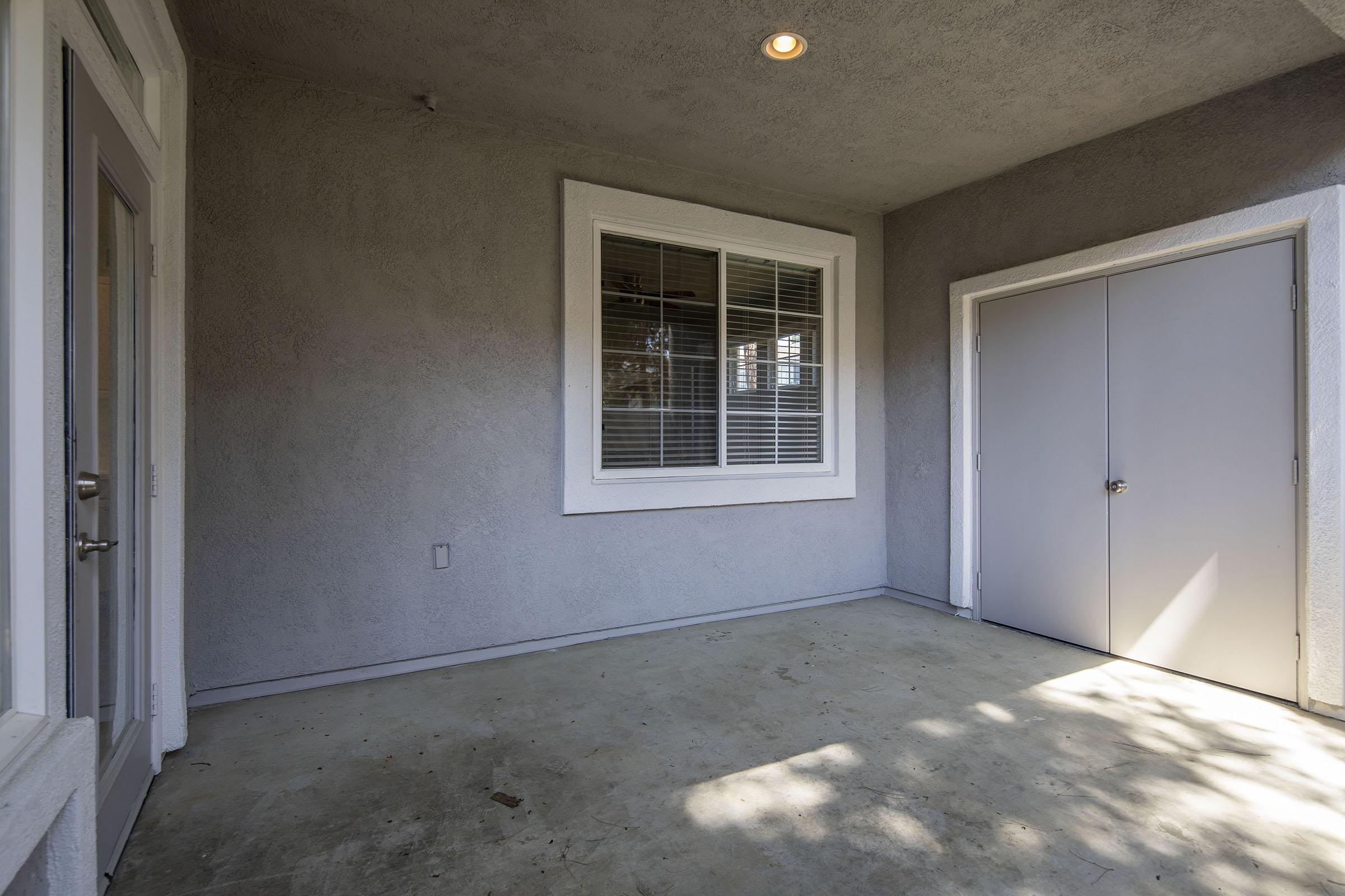 An empty porch or covered entryway with gray walls, a window with white blinds, and a door on the right. The floor is bare concrete, and there is a ceiling light fixture above. The space is well-lit with natural light coming from the window.