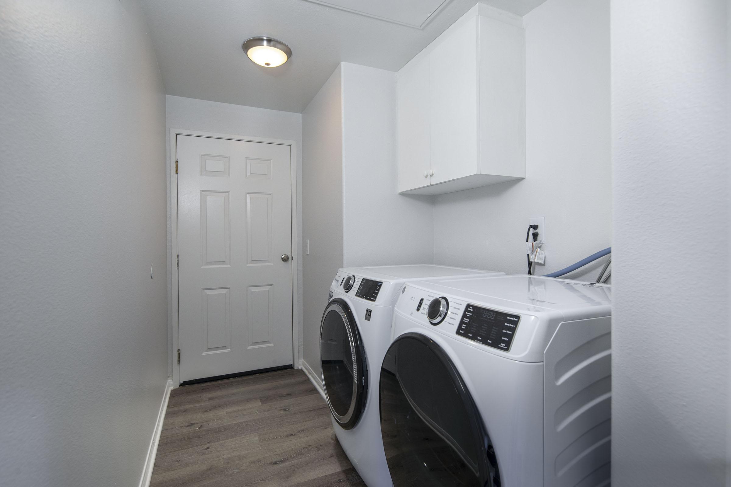 A modern laundry room featuring a pair of front-loading washing machines and a matching dryer. The appliances are positioned against a light gray wall. Above them, there is a wall-mounted cabinet for storage. The room has a door leading out, and the floor is finished with wood-like laminate flooring.