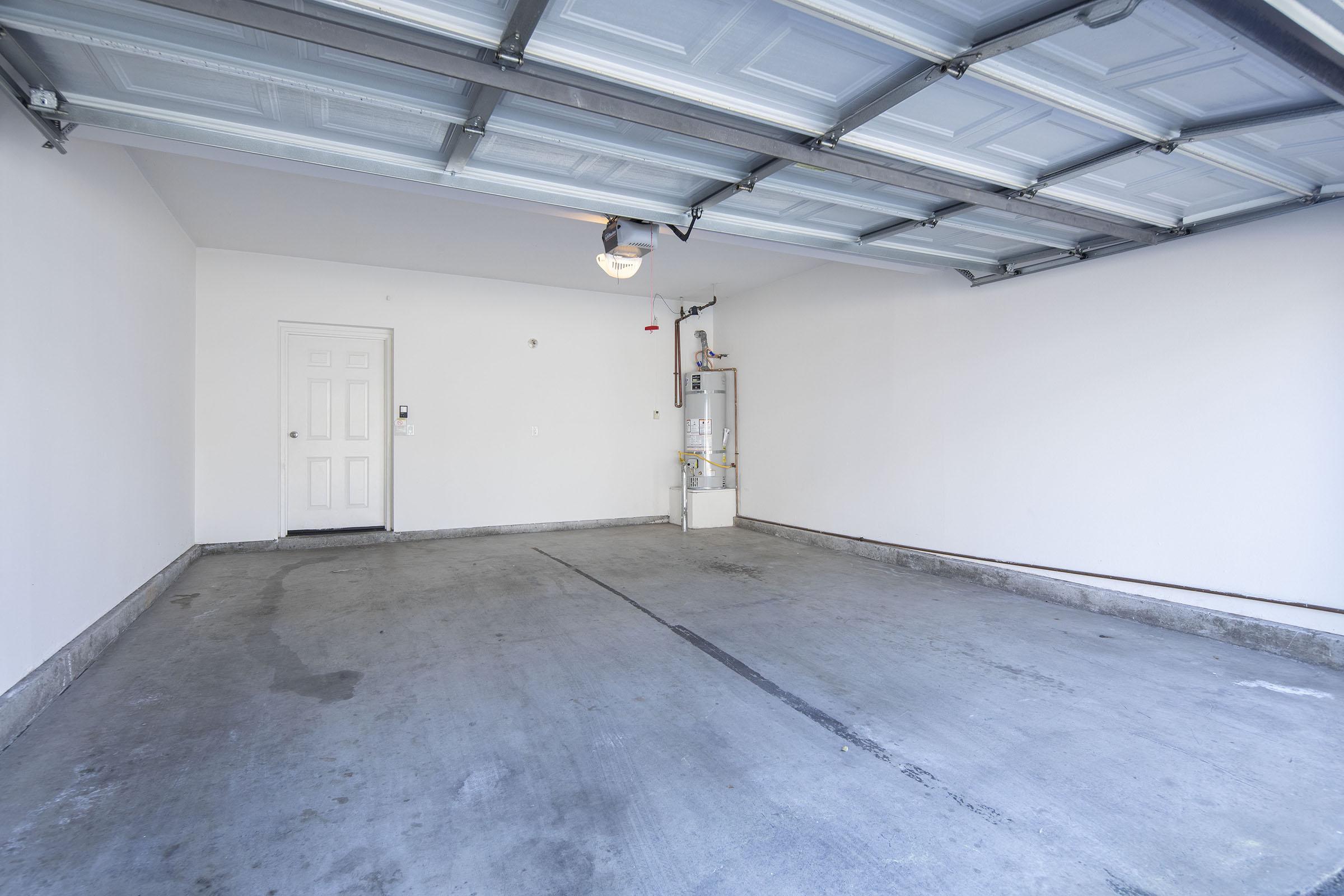 A spacious, empty garage with a concrete floor, white walls, and a ceiling with overhead lighting. A door leads to another room, and there is a water heater against one wall. The garage door is raised, allowing light to enter.