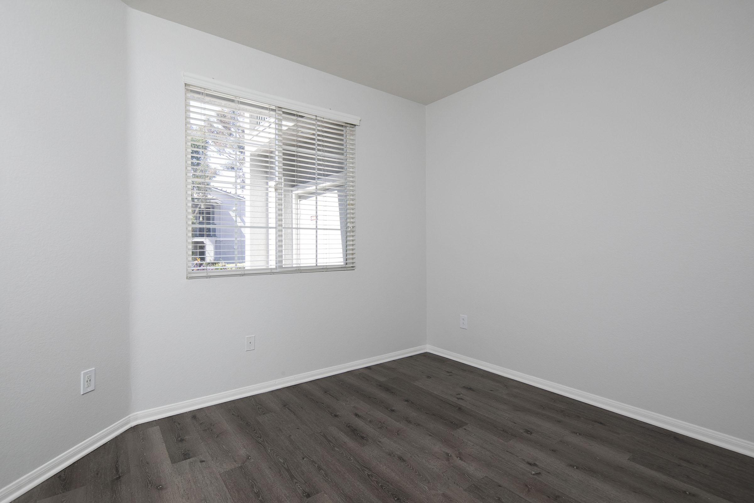 A partially empty room with light gray walls and a wooden floor. There is a large window with closed blinds on one wall, allowing natural light to enter. The room is unadorned, creating a minimalistic and spacious appearance.