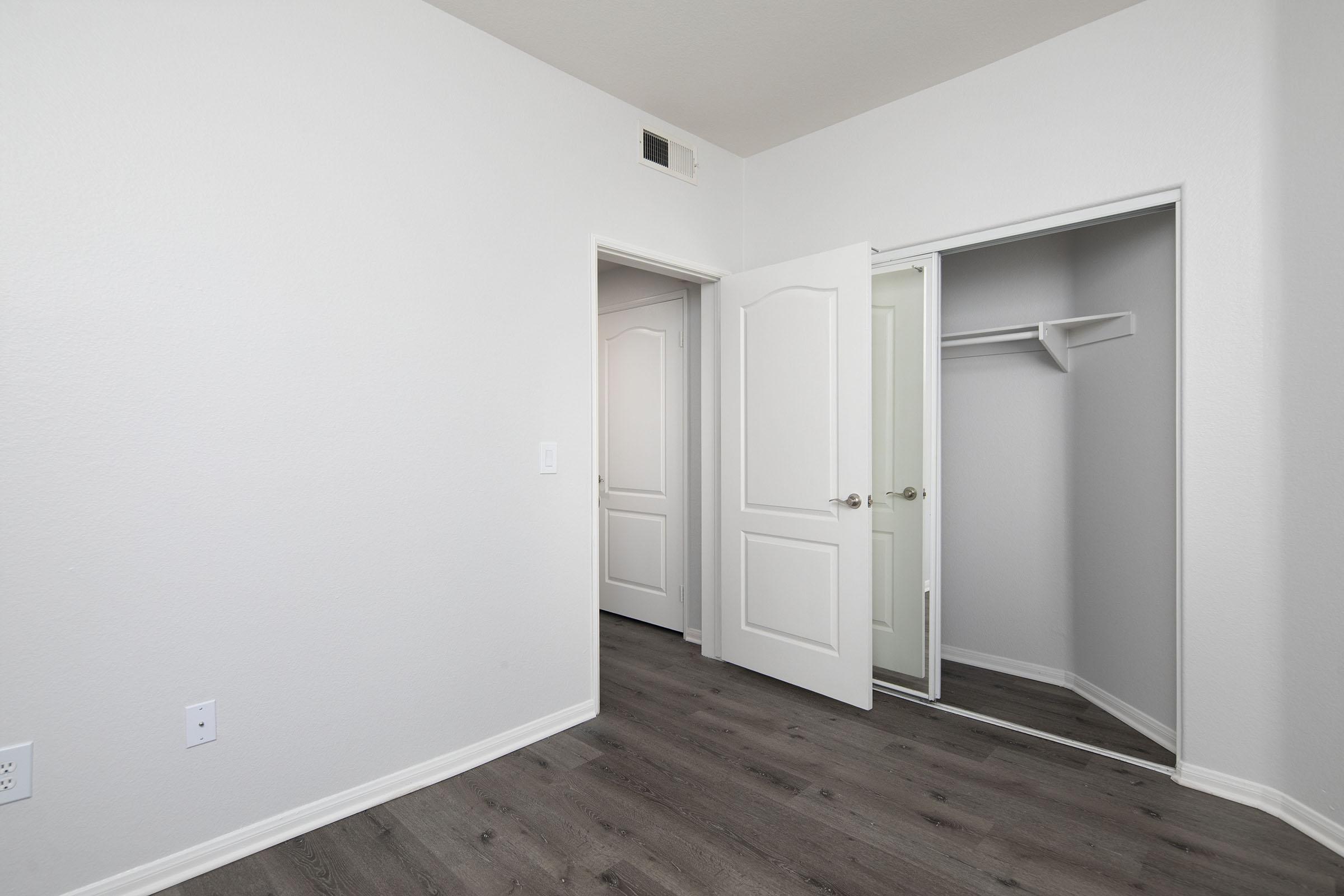 Empty room with light-colored walls and wooden flooring. A double door leads to another room, while a single door opens to a small closet with an open shelf. Natural light fills the space, creating a bright and airy atmosphere.