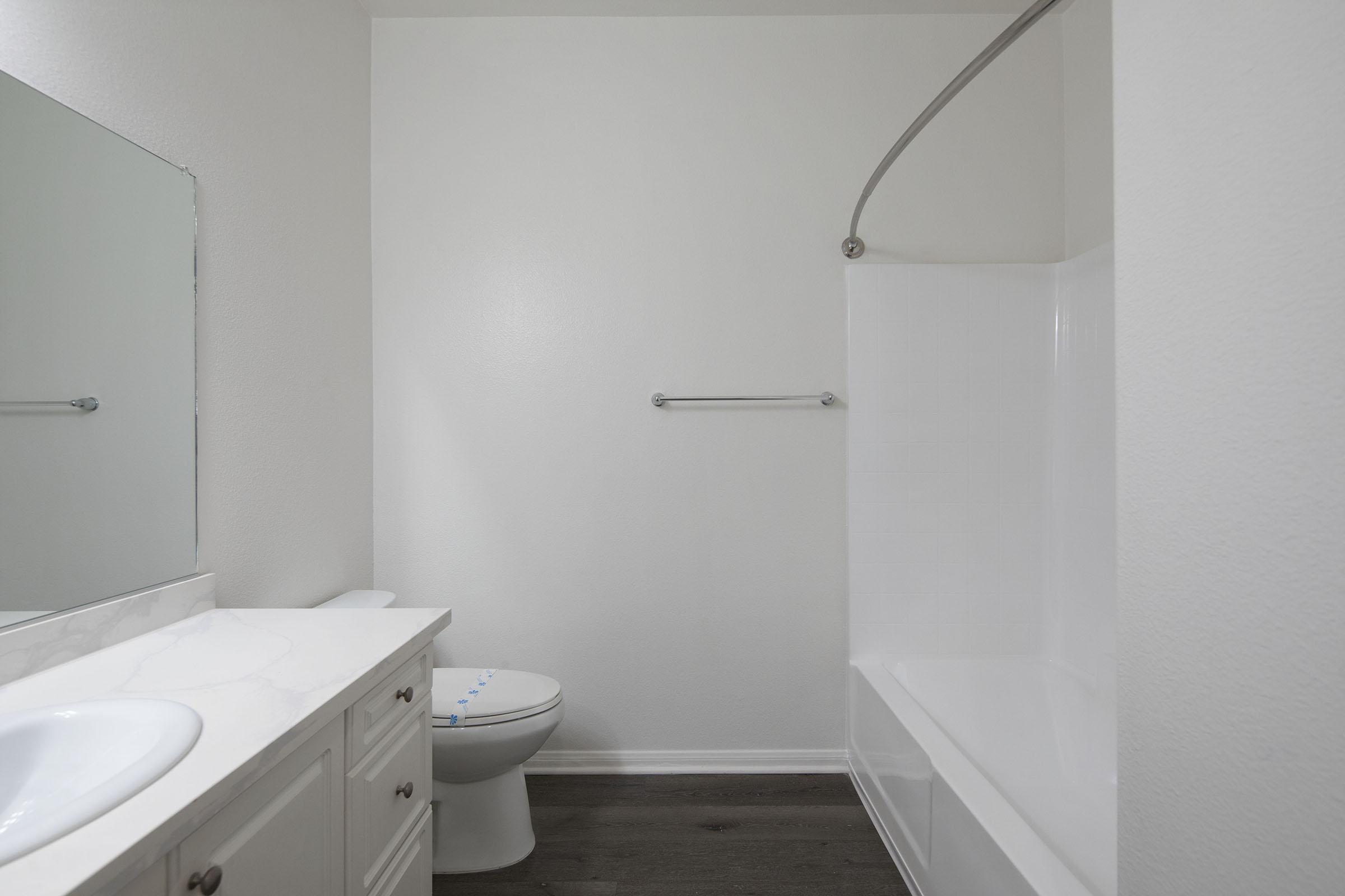 A clean, modern bathroom featuring a white tub and shower, a single sink vanity with a mirror above, and light-colored walls. The bathroom is well-lit and has dark wood flooring, creating a spacious and fresh atmosphere.