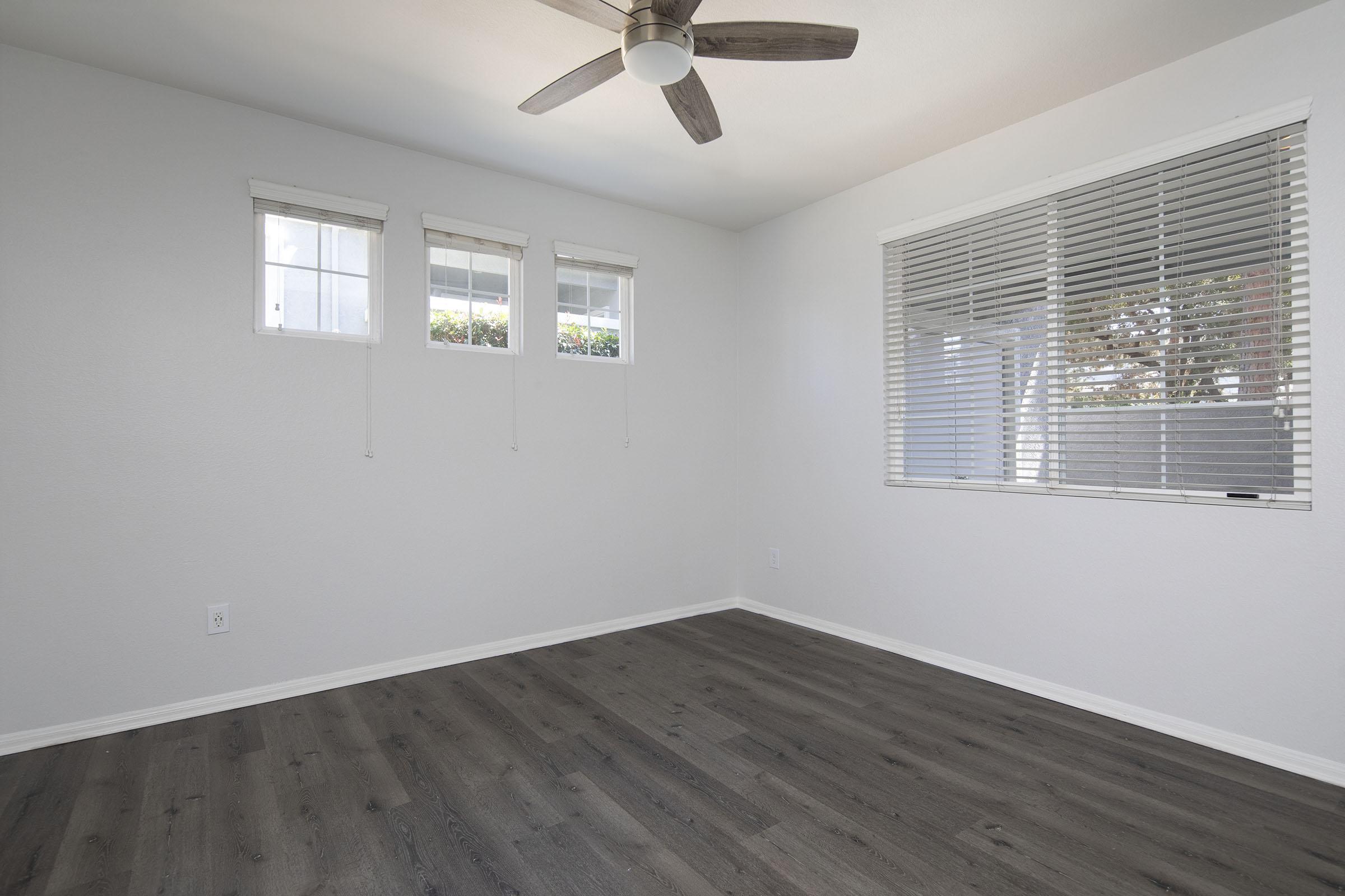 Empty room with light gray walls and dark wooden flooring. Three small windows with white blinds are on one wall, allowing natural light to enter. A ceiling fan is mounted in the center of the ceiling, adding a modern touch to the minimalist space.