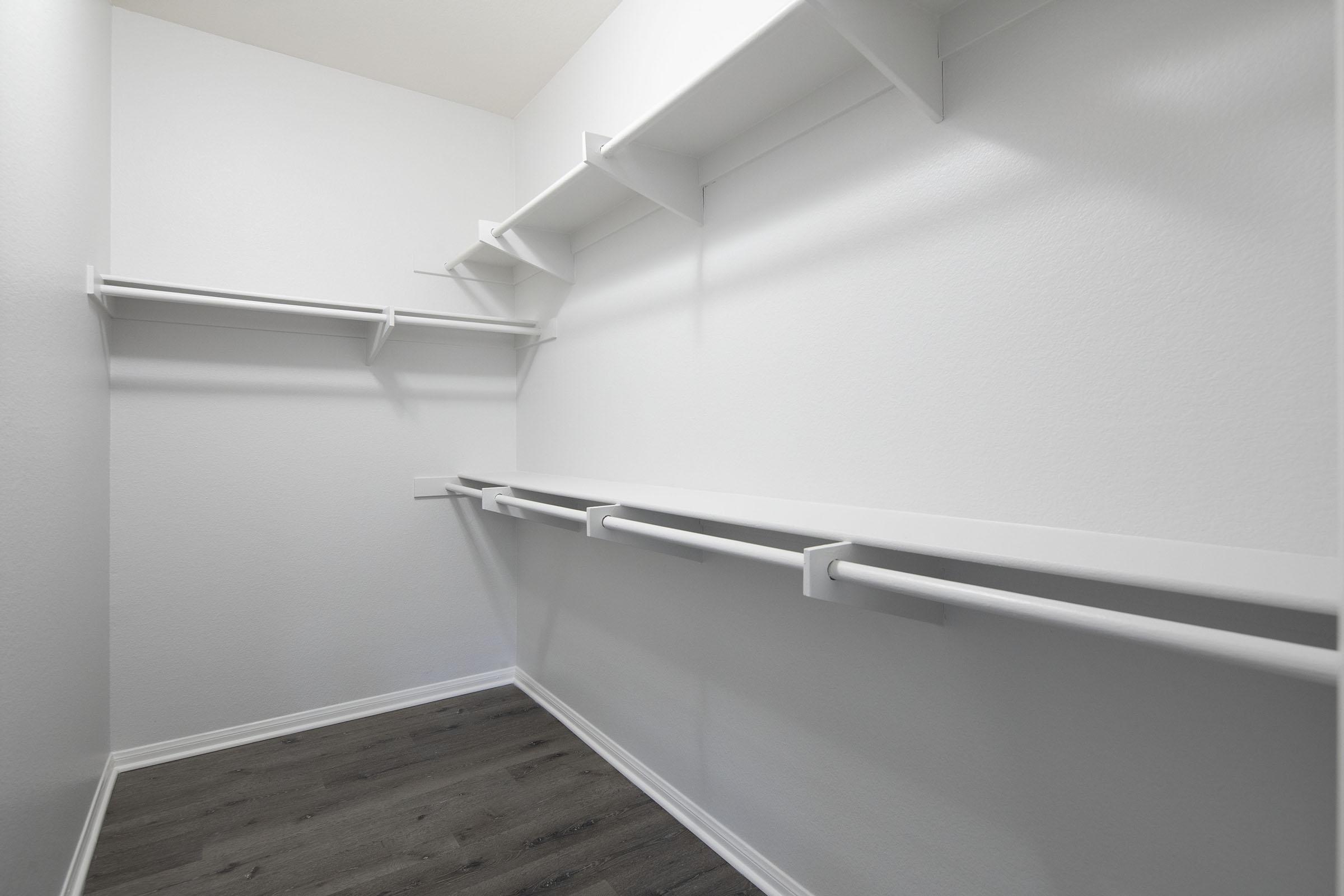 A clean, empty closet featuring white walls and wooden flooring. It has several white shelves mounted on the walls and two horizontal rods for hanging clothes, creating a minimalist storage space. The room is well-lit and organized, emphasizing its spaciousness.