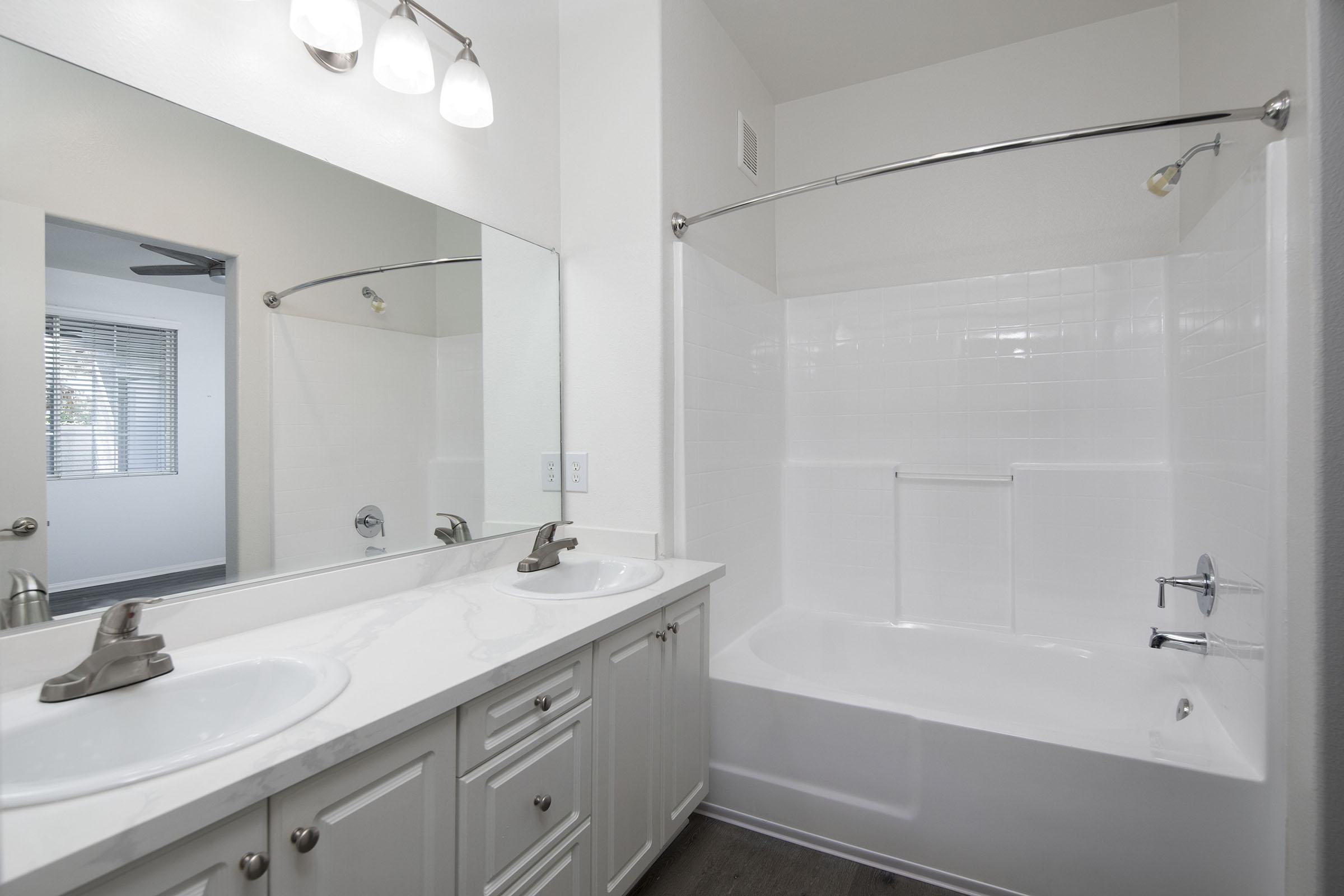 A modern bathroom featuring a double vanity with two sinks and a large mirror above. There is a white bathtub with a curved shower rod and a transparent shower curtain. The walls are painted in a light color, and the floor has dark wood-like tiles. Natural light enters from a nearby window.