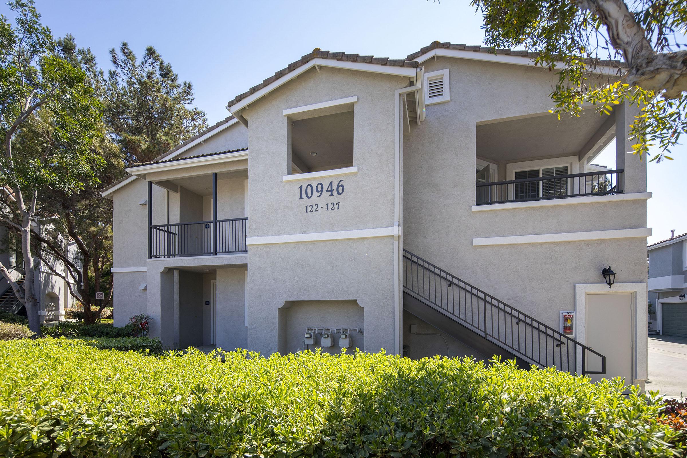 Exterior view of a two-story residential building with a light gray stucco finish. The building has a staircase leading to the upper level, with small balconies visible. Lush green shrubs surround the base of the building, and the numbers "10946" are prominently displayed on the wall. Bright blue sky in the background.