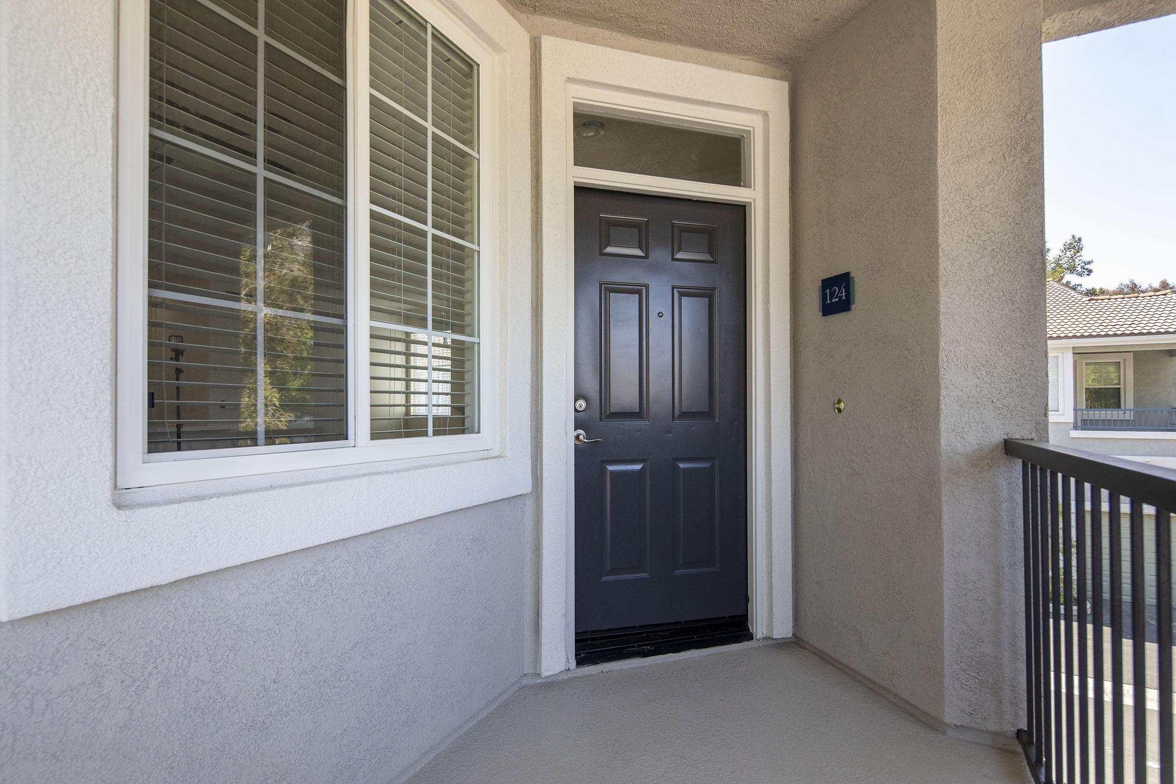 A view of a modern apartment balcony featuring a dark front door with the number 124 on a blue plaque. To the left, there's a window with white shutters and a view of the outside. The floor is light-colored, and the surrounding walls are painted in a neutral tone.