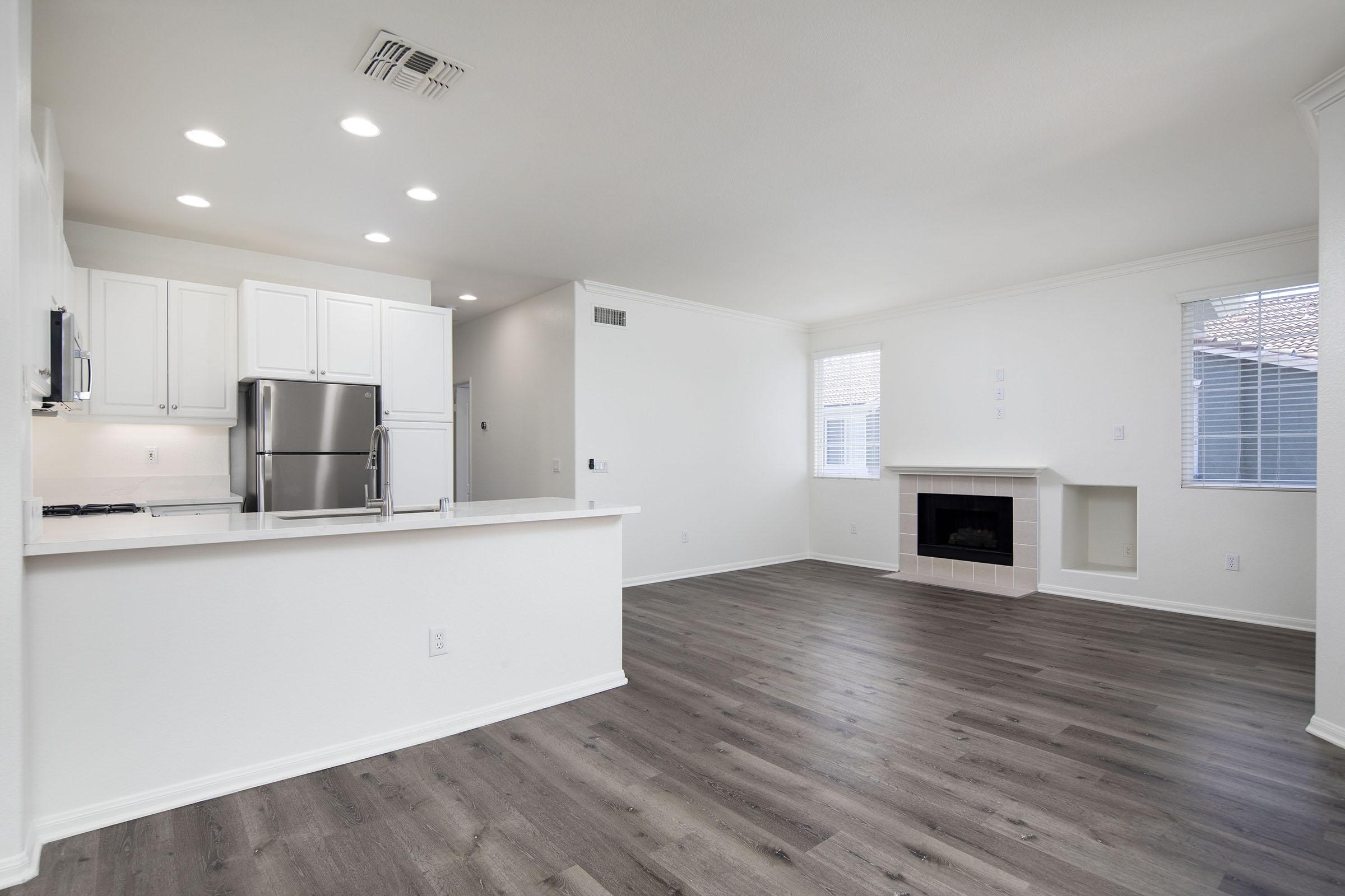 Interior view of a modern, open-concept living space featuring a kitchen with white cabinets and stainless steel appliances. The living area has a fireplace and large windows, allowing natural light to fill the room. The flooring is a light wood finish, creating a warm and inviting atmosphere.