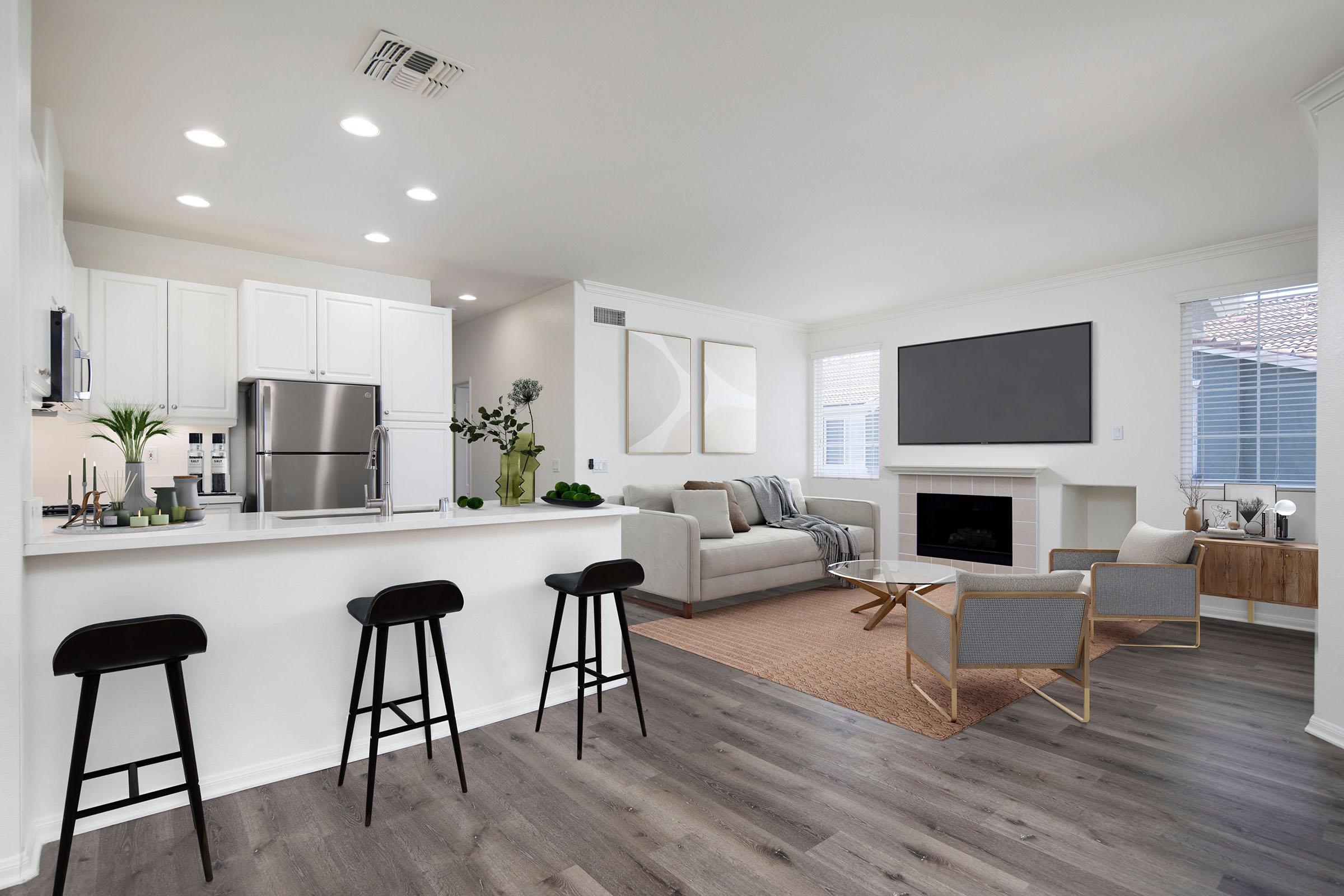 Modern living room and kitchen area in an open-concept design. Features a light-colored sofa, gray armchairs, a wooden coffee table, and an entertainment unit with a flat-screen TV. The kitchen includes white cabinets and stainless steel appliances, with three black stools at the breakfast bar. Large windows provide natural light.