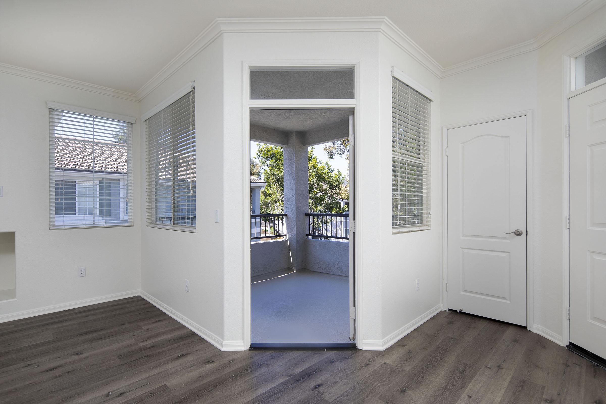 A bright, airy room with neutral-colored walls and wooden flooring. Two large windows on the left allow natural light, while a sliding door in the center leads to a small balcony. The door on the right is closed, and the overall space has a modern, inviting feel.