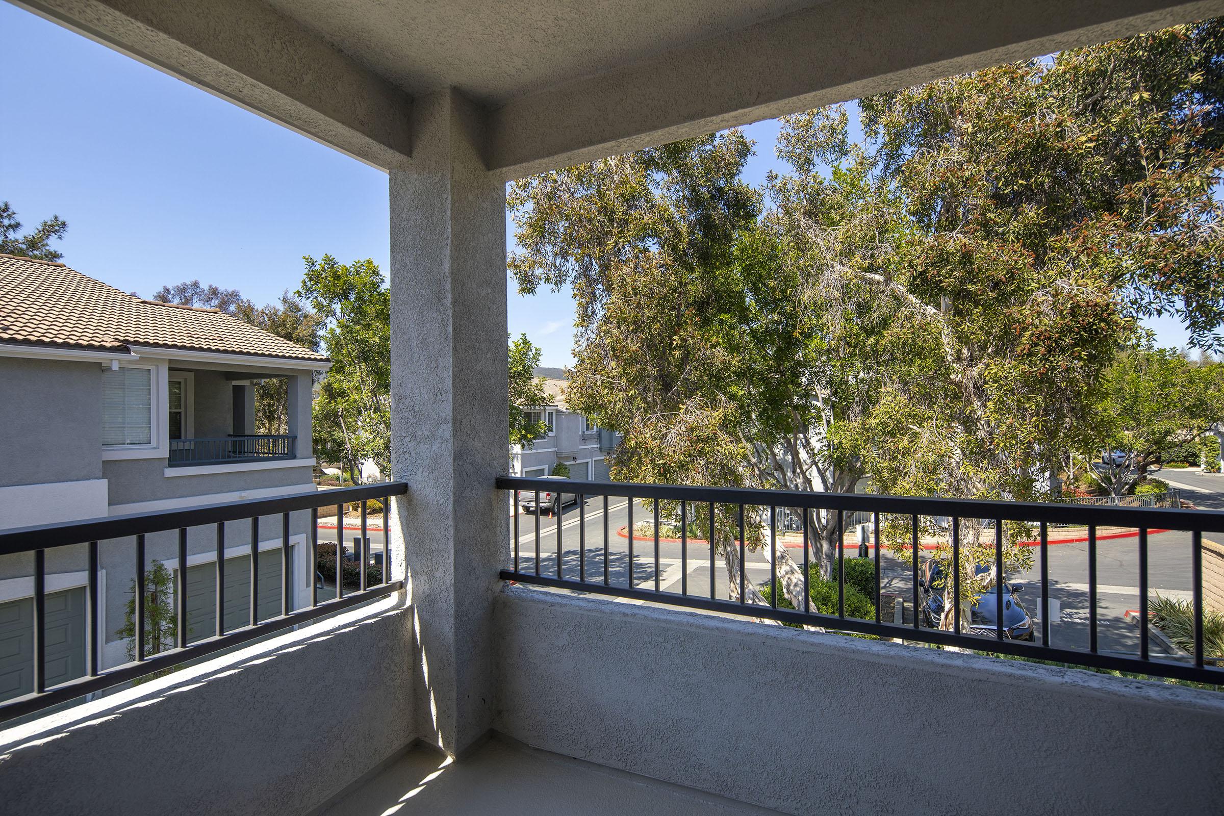 View from a balcony showcasing a residential area with two-story buildings, lush green trees, and a clear blue sky. The scene conveys a peaceful atmosphere with sunlight streaming in.