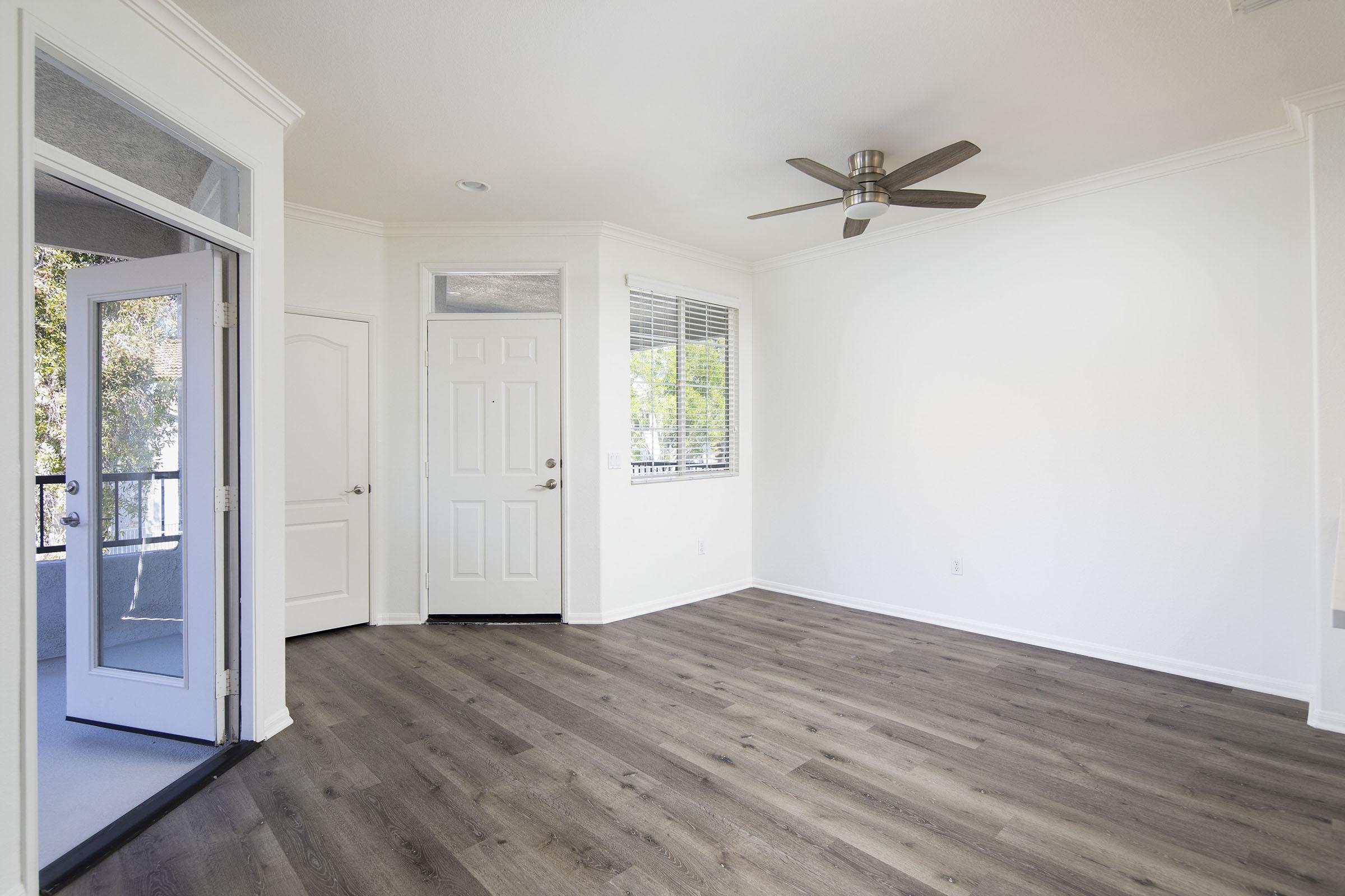 A bright, empty room featuring light wooden flooring, white walls, and a ceiling fan. Two doors are visible: a closed white door and an open door leading outside. Large windows allow natural light to fill the space, creating a spacious and airy atmosphere.