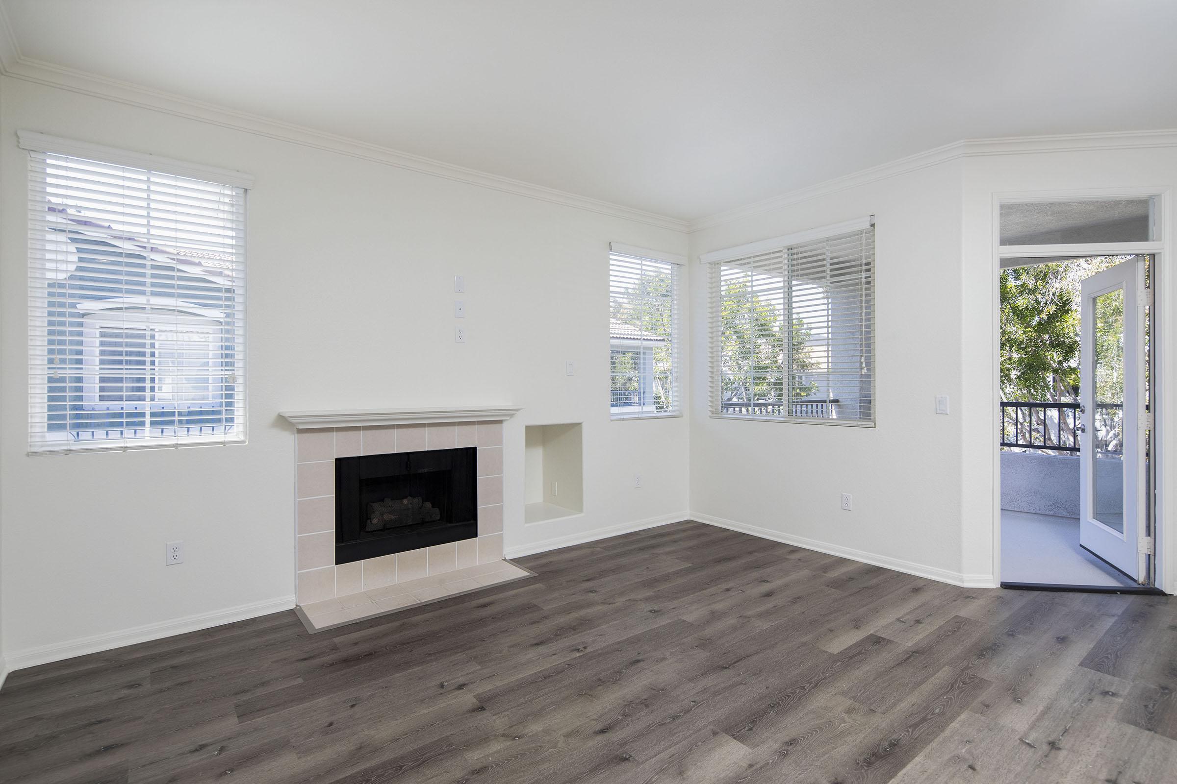 Spacious, empty living room featuring a fireplace with a cream-colored surround, large windows with blinds, and light-colored walls. It has a modern look with wood-style flooring and an entrance leading to a balcony area. Natural light enhances the bright and airy atmosphere.