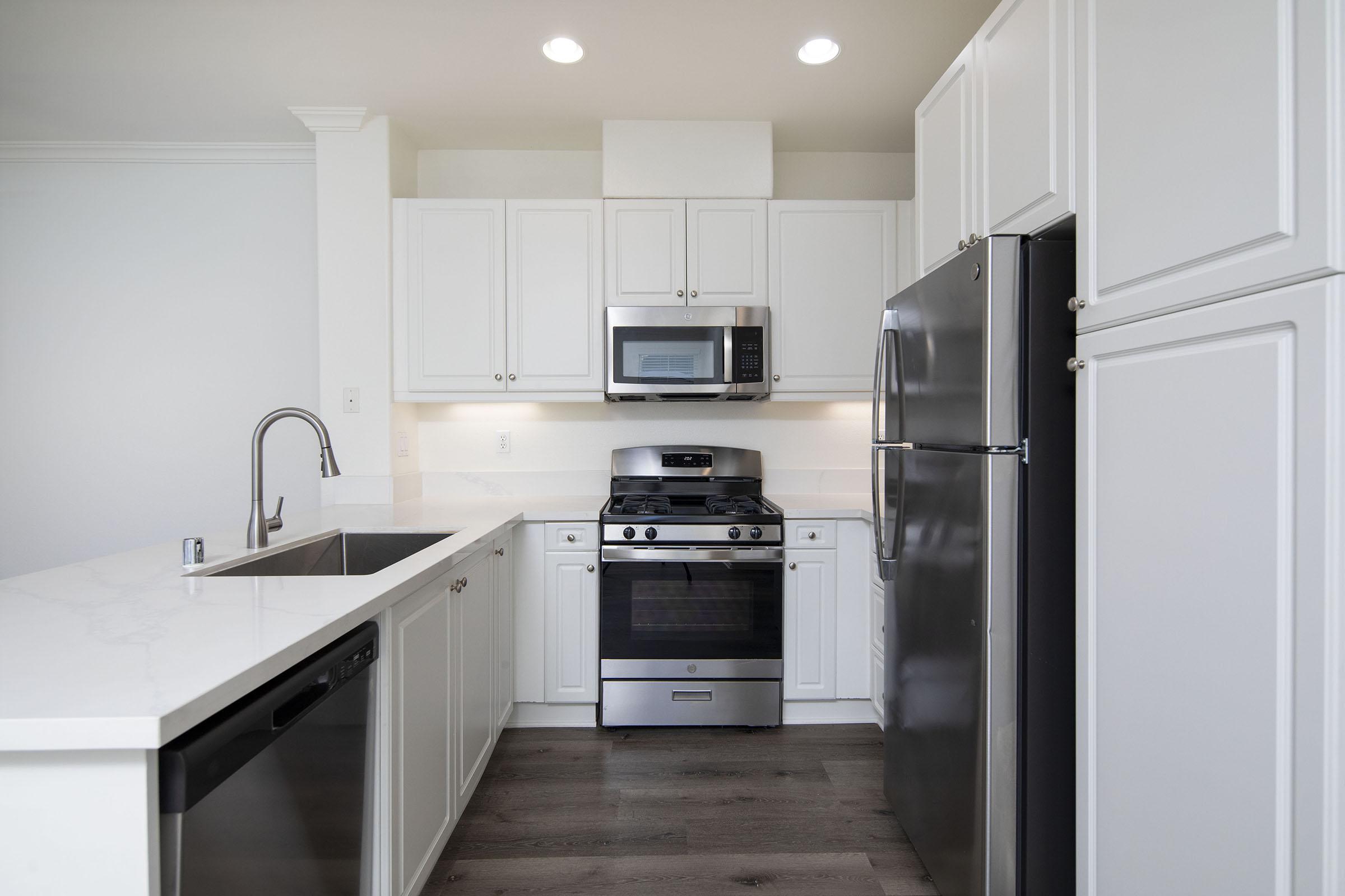 A modern kitchen featuring white cabinetry, stainless steel appliances including a microwave, gas stove, and refrigerator. The countertop is a light-colored stone, complemented by a double basin sink. The floor is covered in light wood-like planks, and the room is well-lit with overhead lighting.