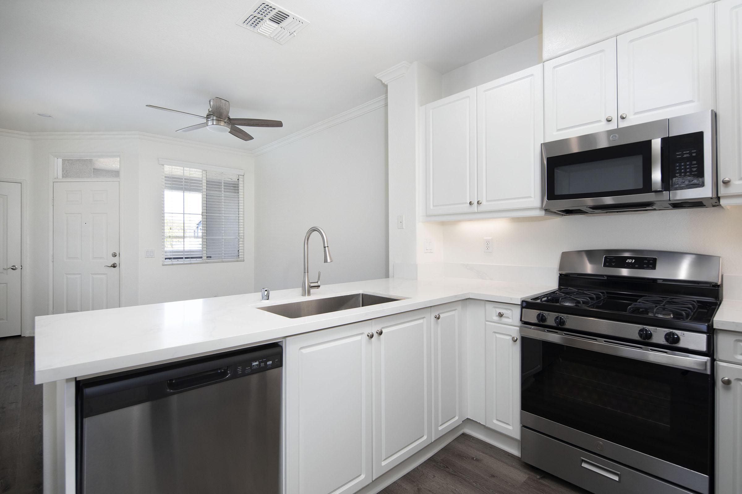 Modern kitchen featuring white cabinetry, stainless steel appliances including a microwave and a gas stove, a dishwasher, and a sleek sink. The countertop is made of light-colored stone, and there's a ceiling fan above. The room has ample natural light from a nearby window and includes a door leading to another room.