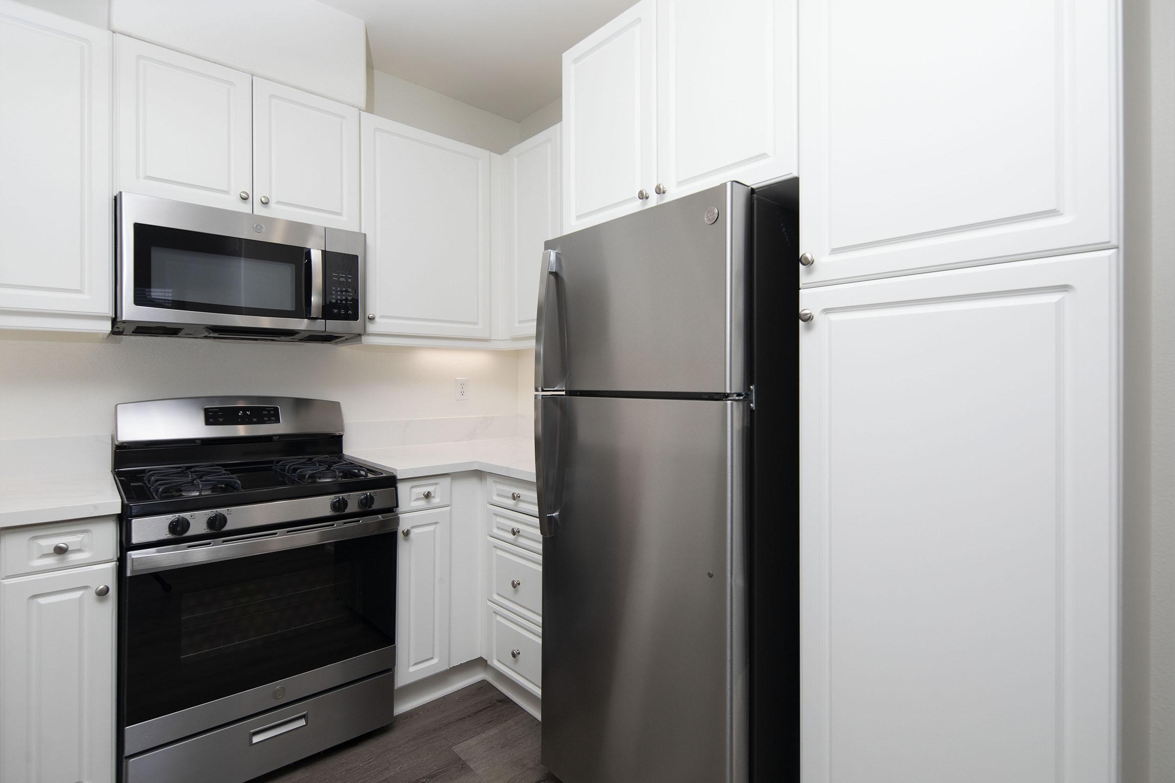 A modern kitchen featuring white cabinets, a stainless steel refrigerator, a gas stove with an oven, and a microwave above the stove. The countertop is light-colored, and the space is well-lit, creating a clean and contemporary look.