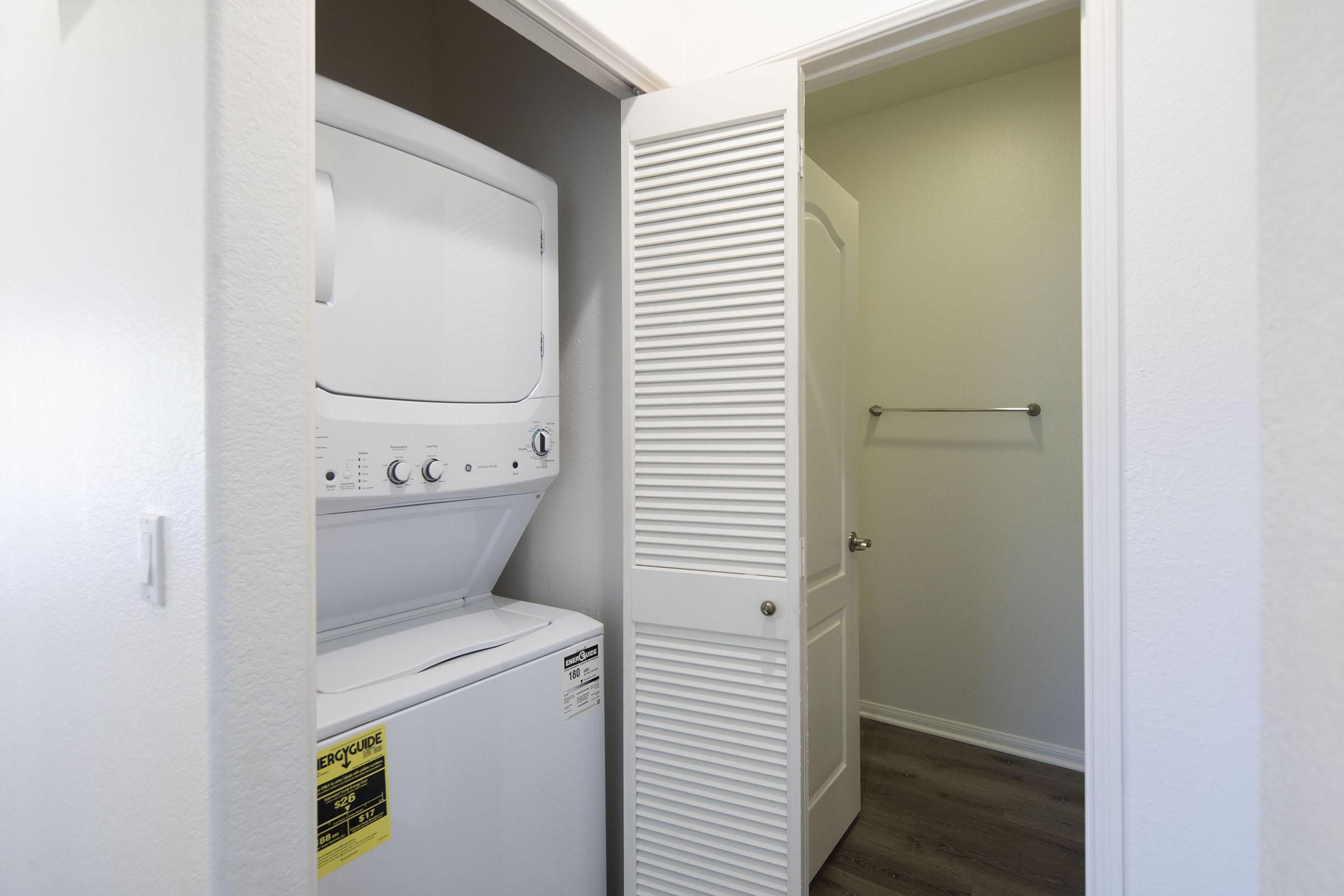 A small laundry area featuring a stacked washer and dryer positioned against a wall. A door partially open reveals a small storage space with a towel bar. The surrounding walls are painted in light colors, and the flooring is a neutral laminate.